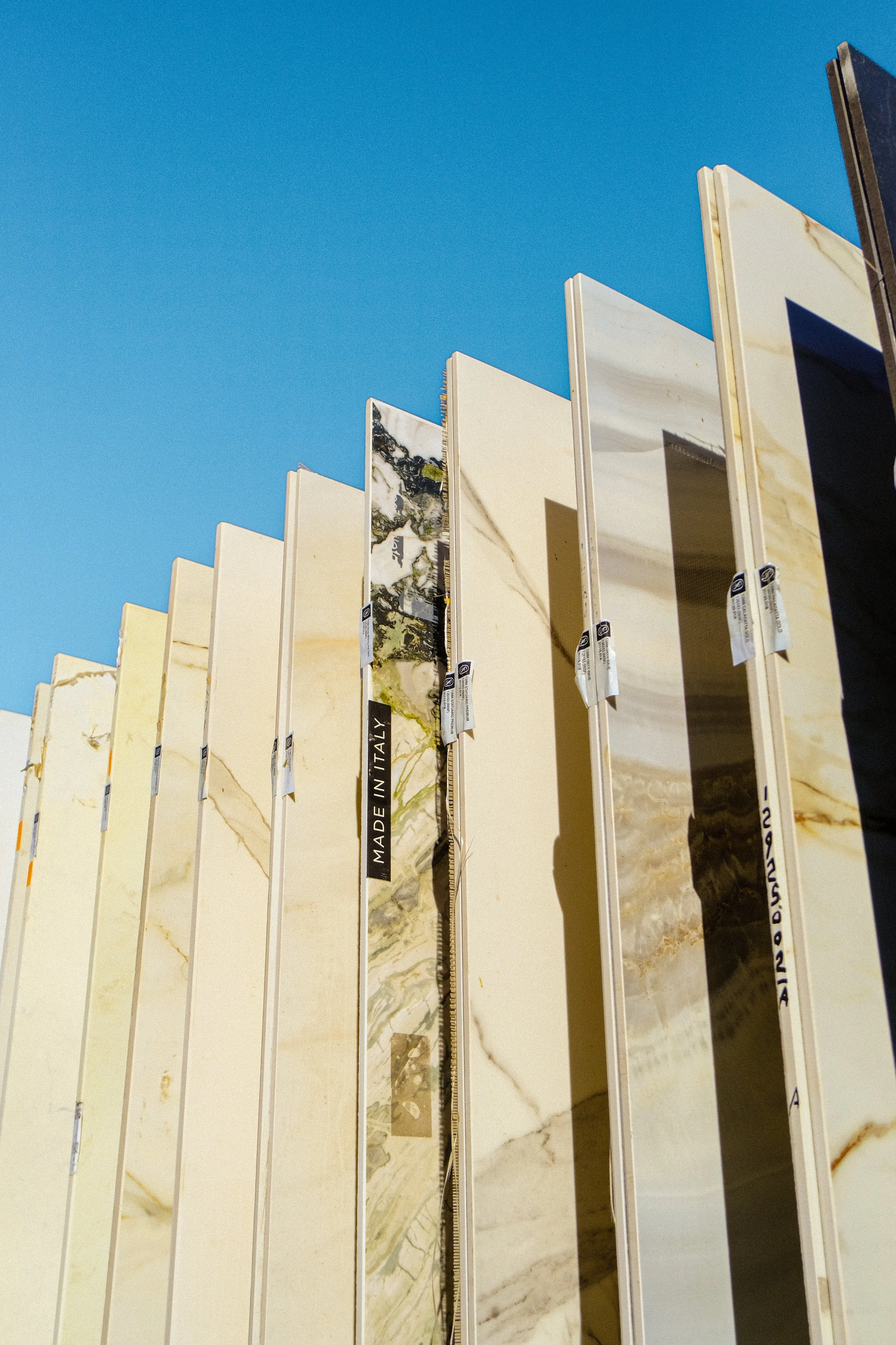 Rows of marble and stone tiles displayed outdoors against a clear blue sky, with labels indicating they are made in Italy.