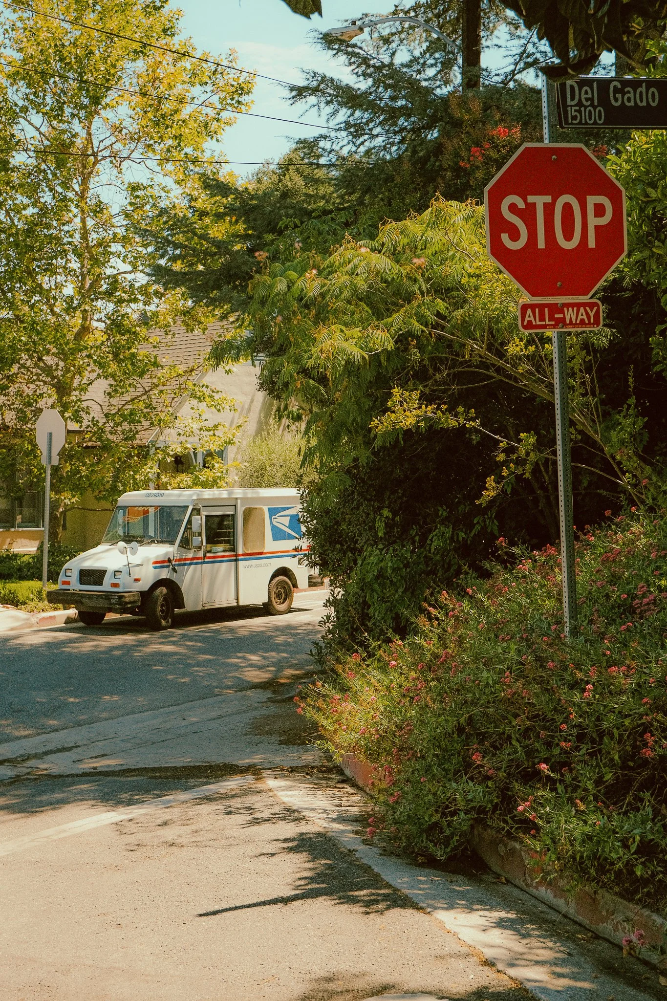 A suburban street corner with a red stop sign and a USPS mail delivery truck parked on the side of the street, surrounded by green trees and bushes, and a street sign indicating Del Gado.