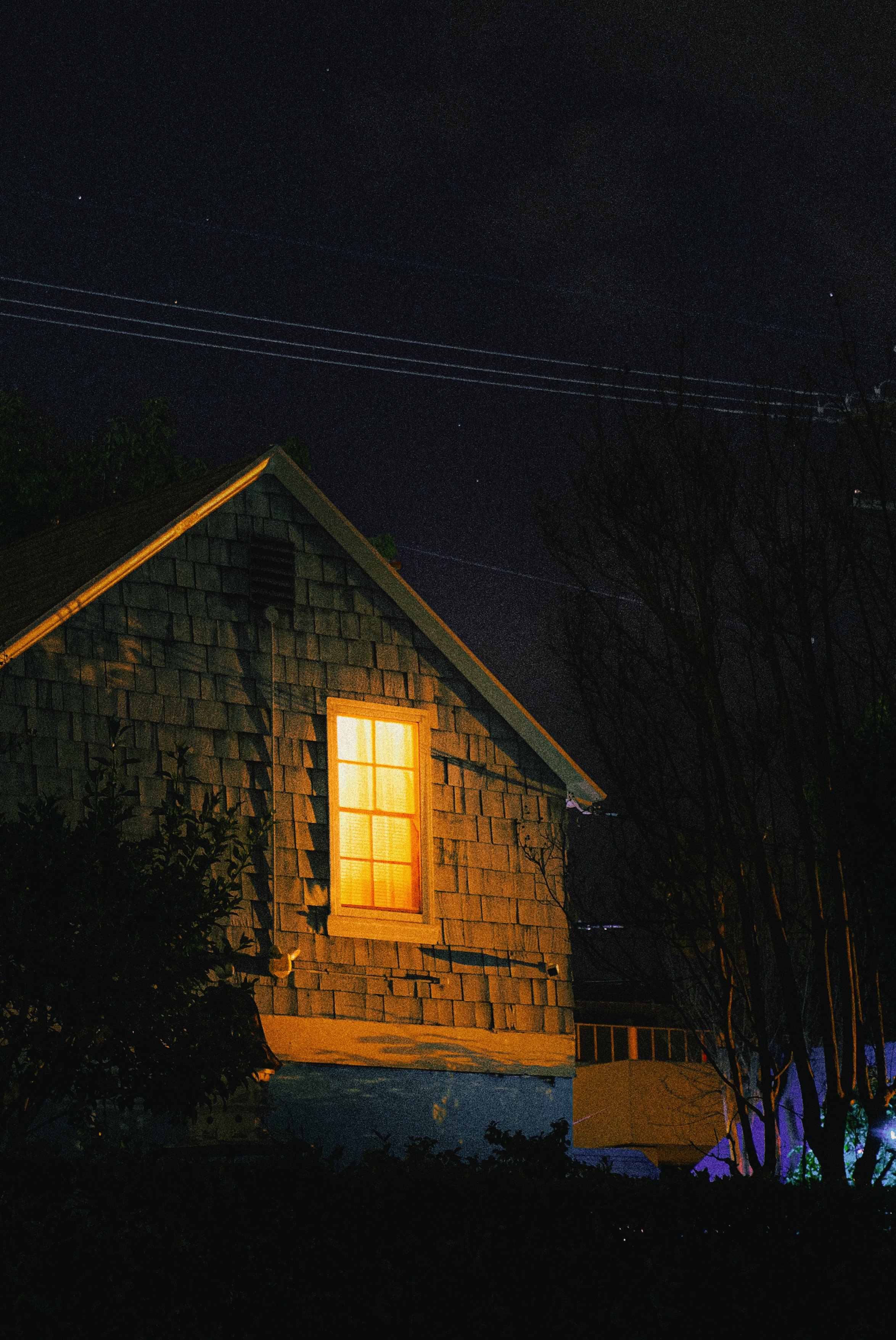 A house with a single illuminated window at night, surrounded by trees and power lines against a starry sky.