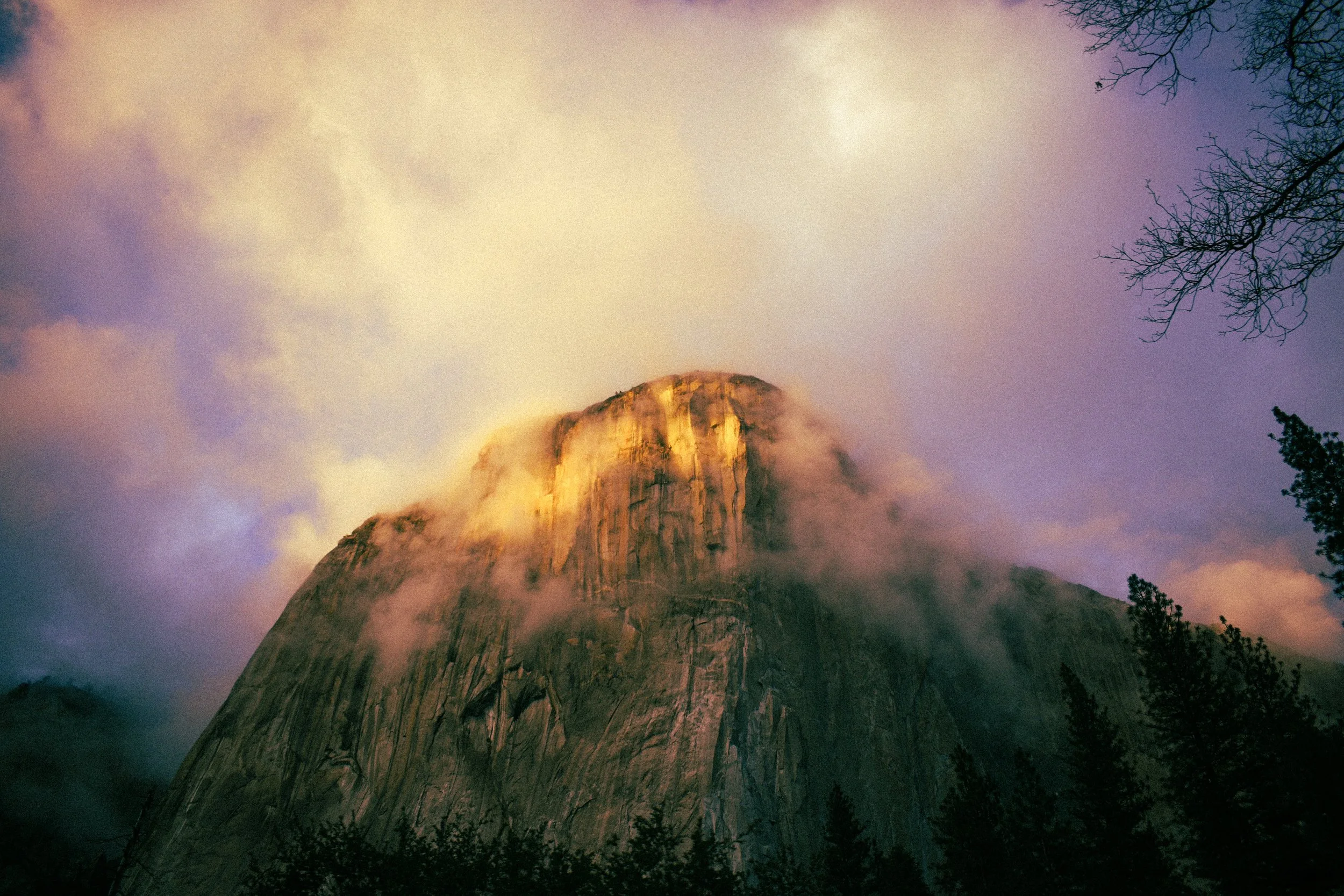 A tall mountain partially covered in clouds during sunset with trees in the foreground.