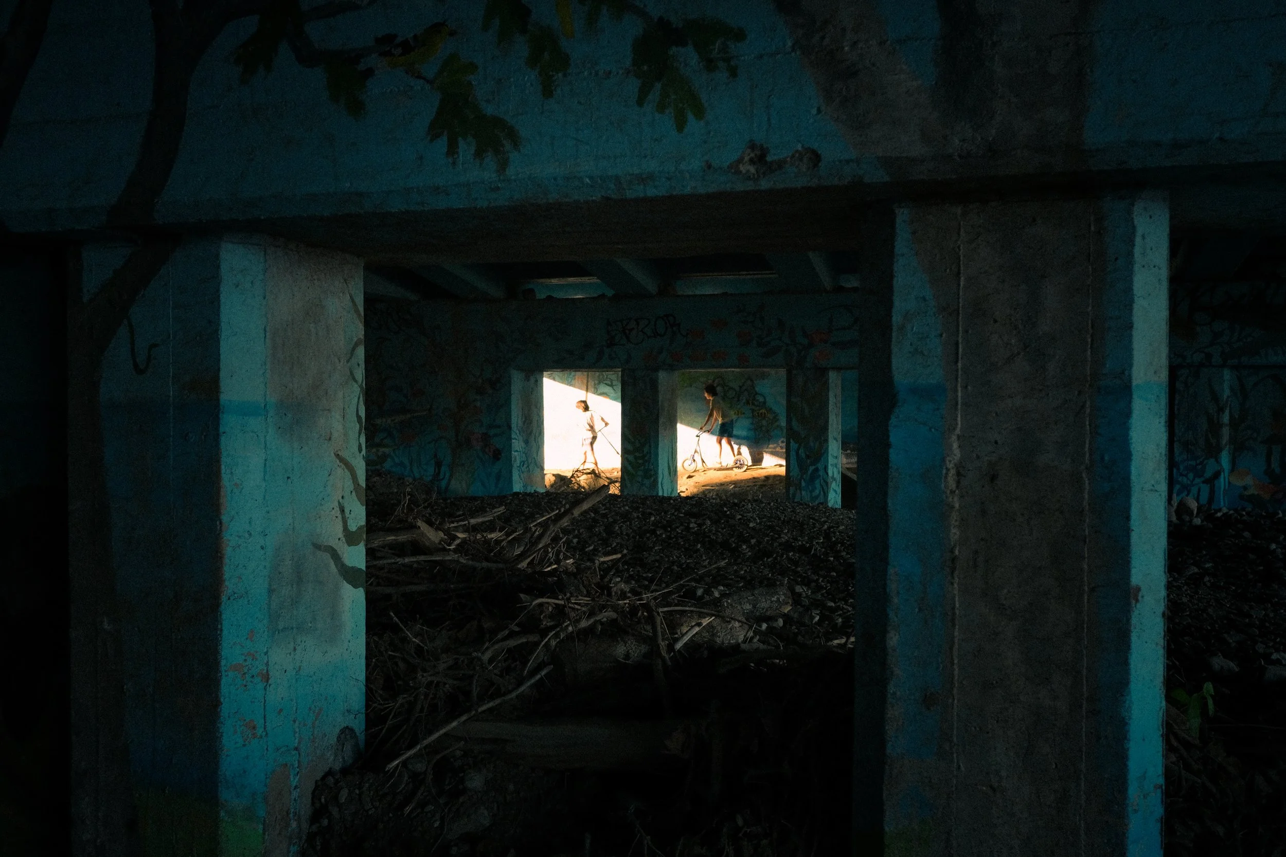 Underneath a bridge or overpass with graffiti-covered concrete pillars, silhouettes of children playing on scooters are visible in the bright light at the far end of the tunnel.