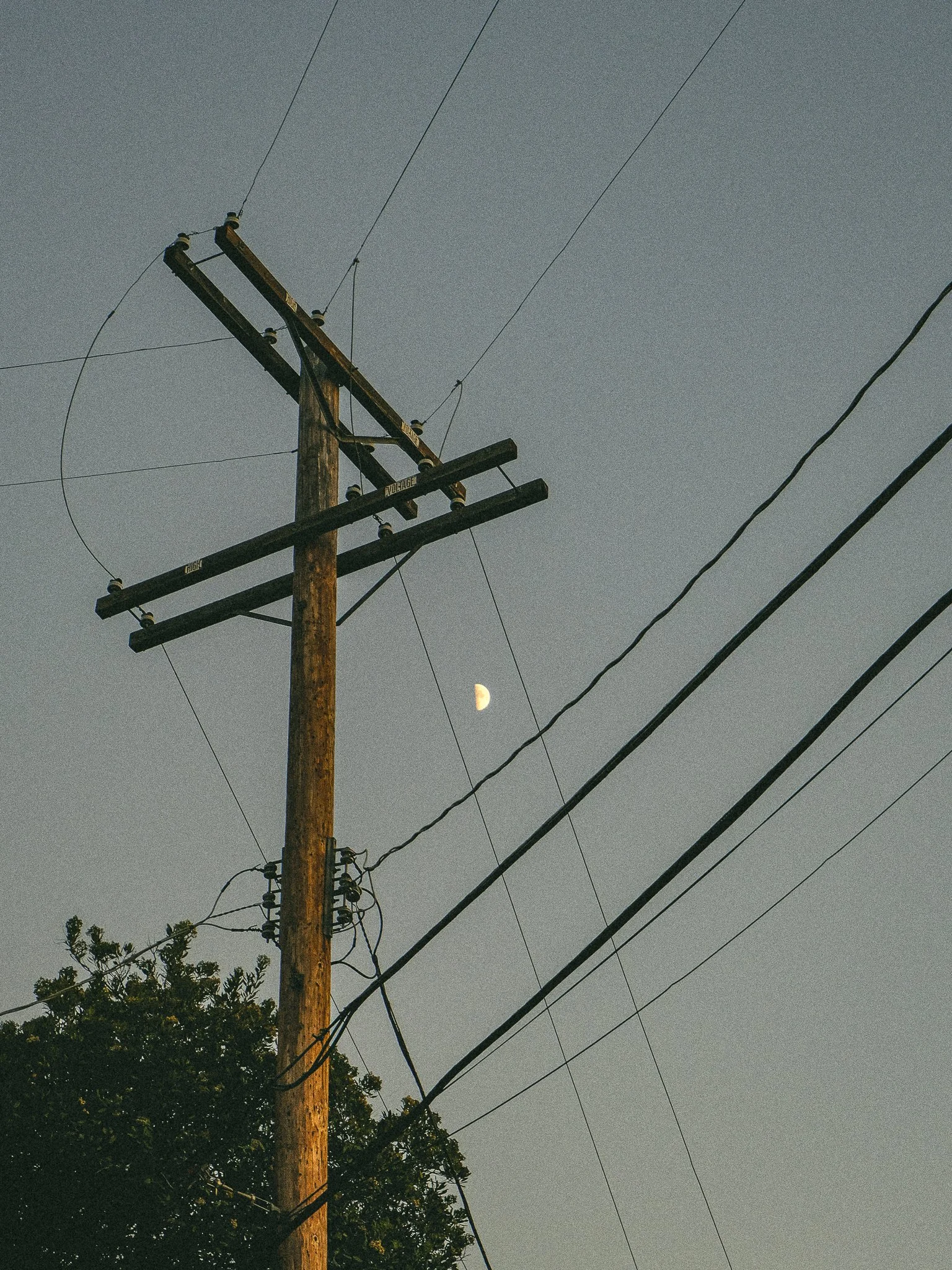 A wooden utility pole with multiple power lines against a clear sky with a visible crescent moon.