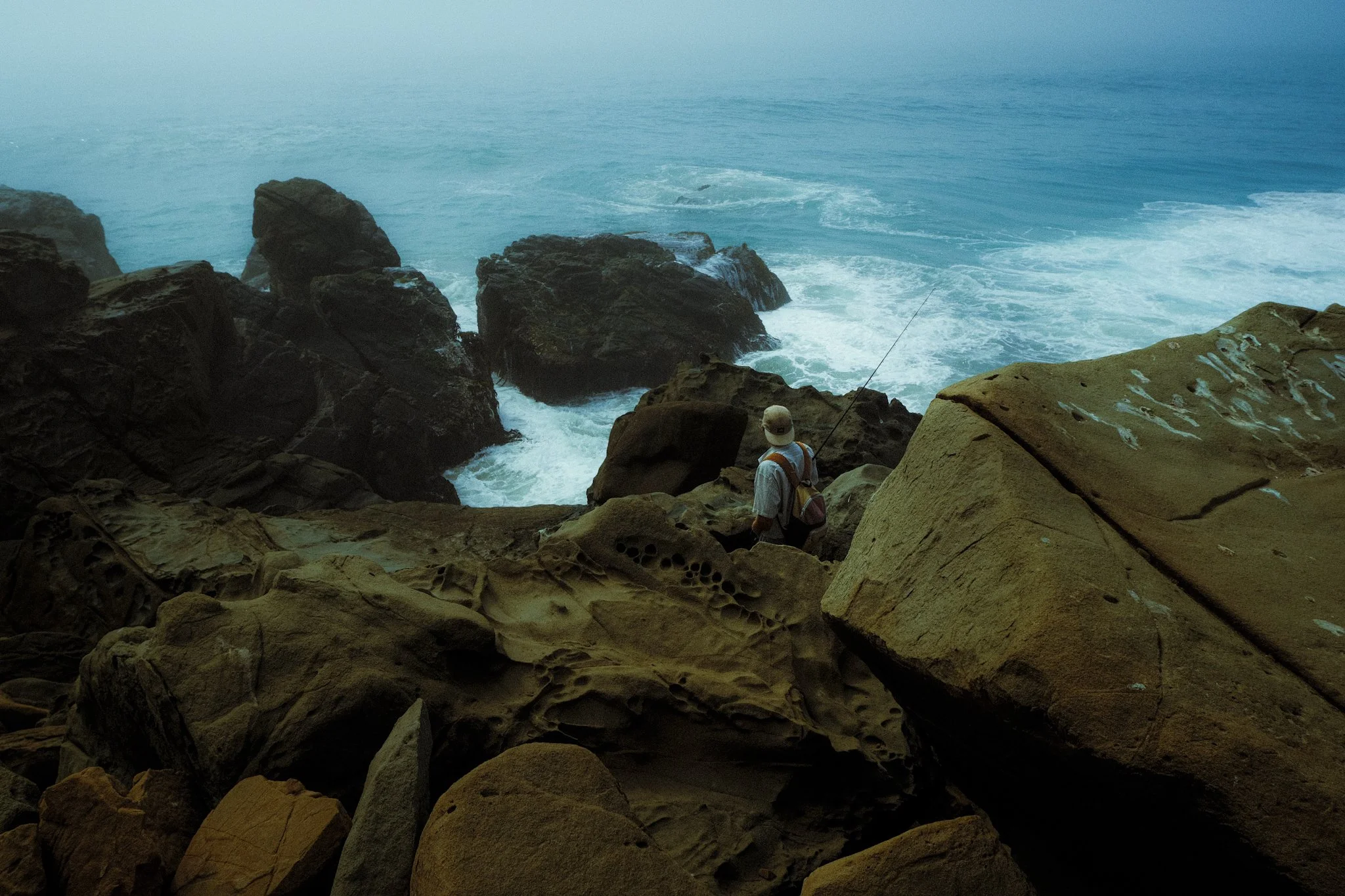 A person fishing on rocky cliffs overlooking the ocean with waves crashing against the rocks.