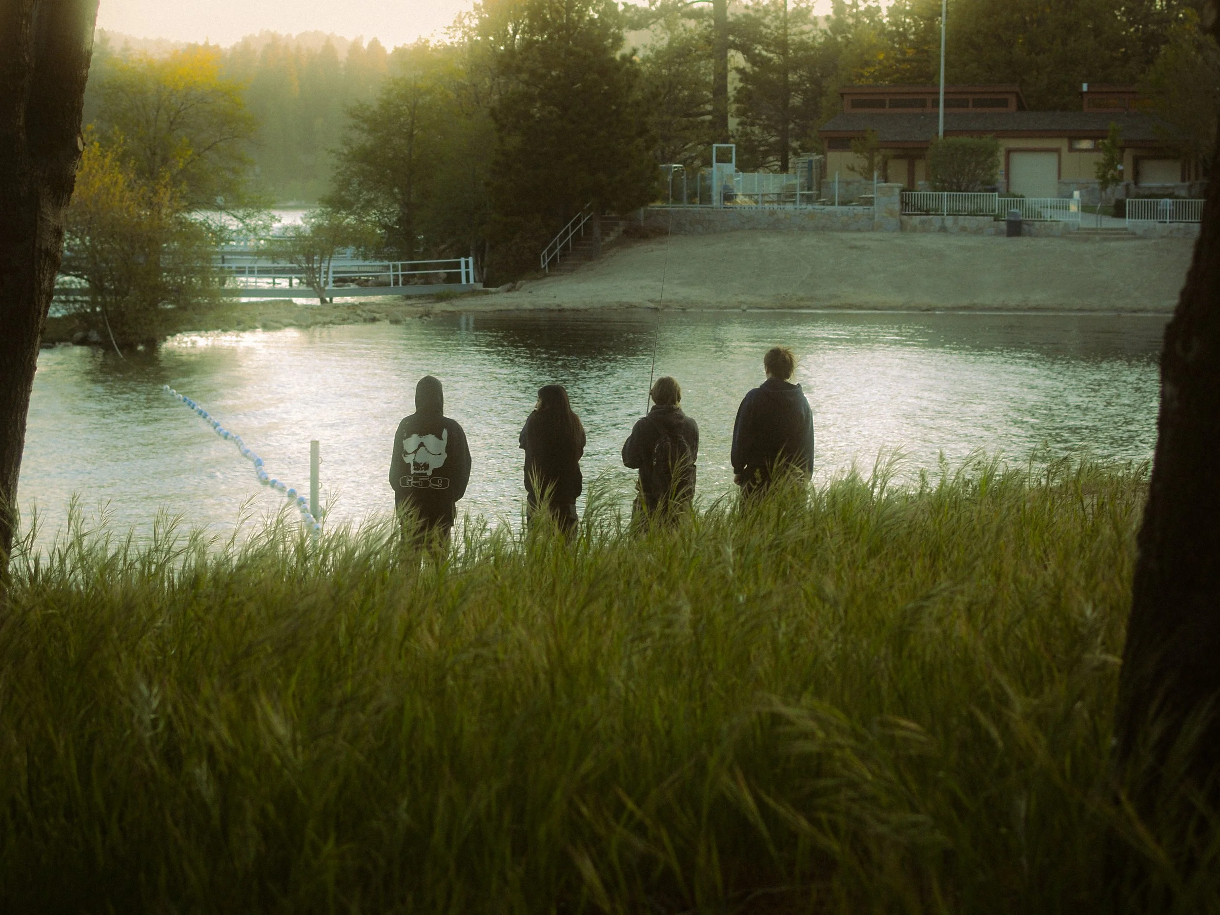 Four people stand on the grassy bank of a lake, with houses and trees in the background, and two trees framing the scene on either side.