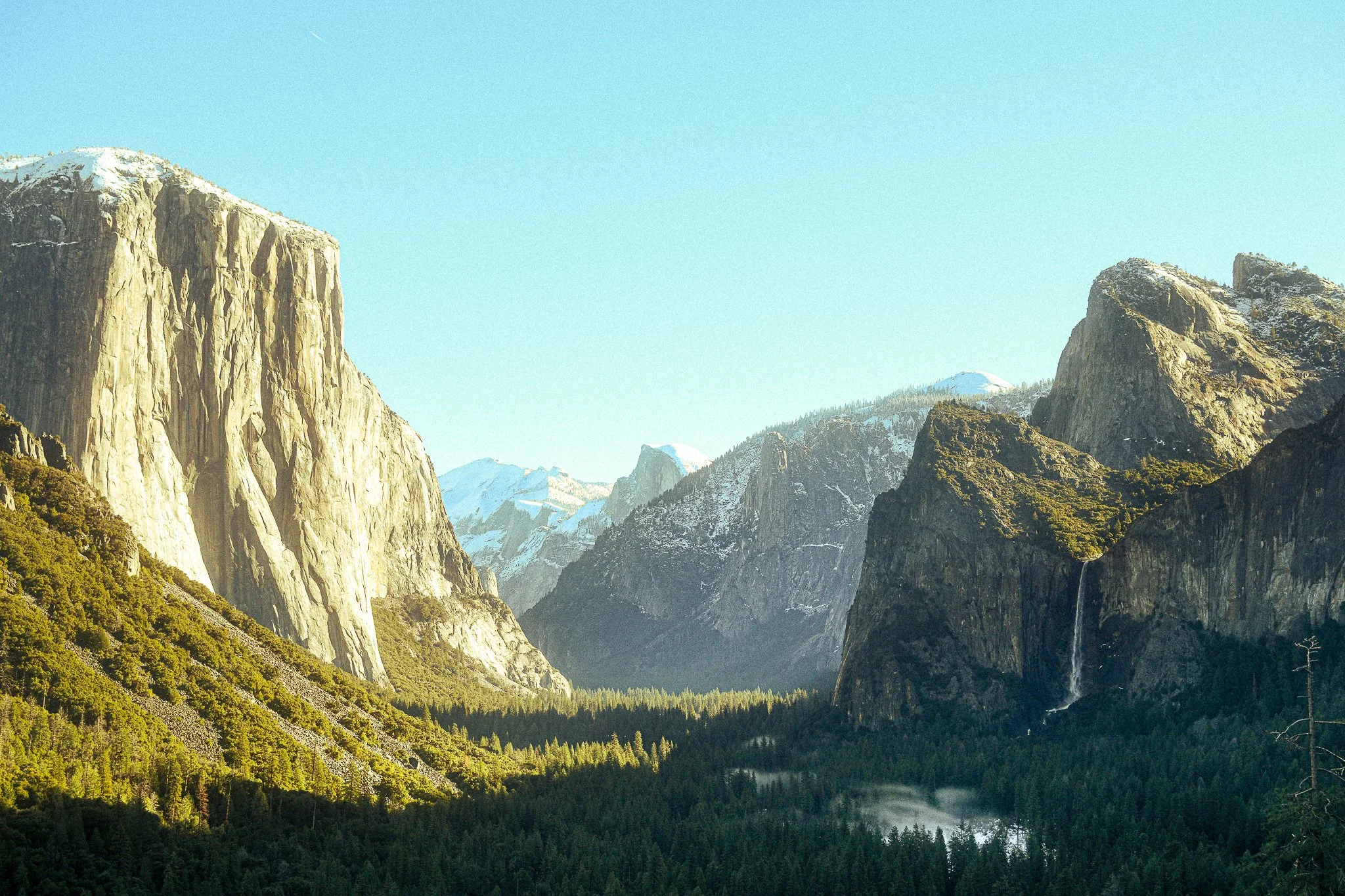 A panoramic view of Yosemite National Park featuring granite cliffs, snow-capped mountains, a forest, and a waterfall.