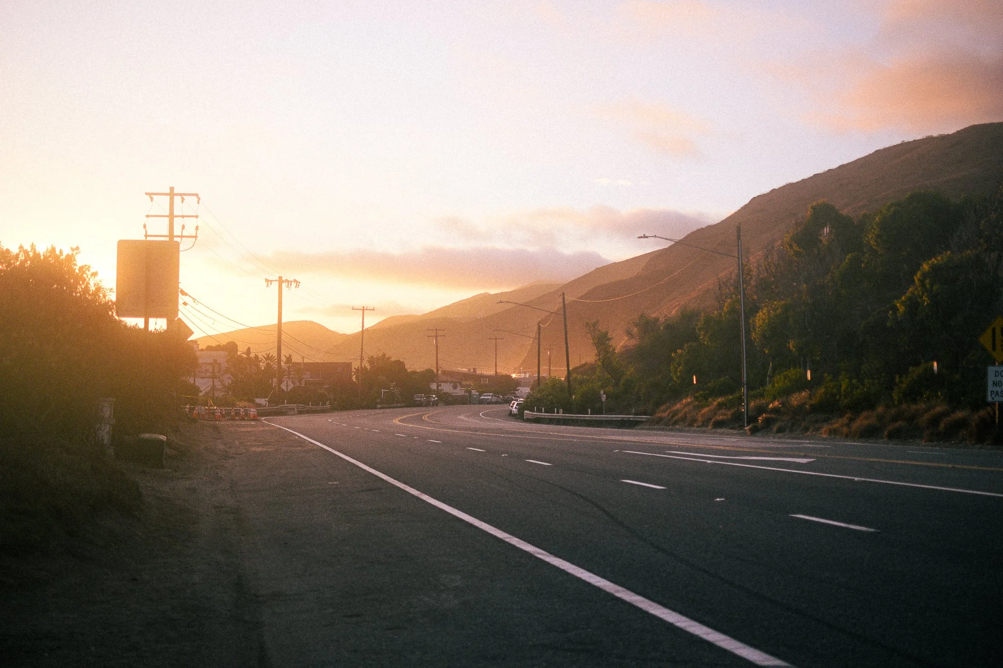 A scenic highway at sunset with mountains in the background, trees along the sides, and power lines running parallel to the road.