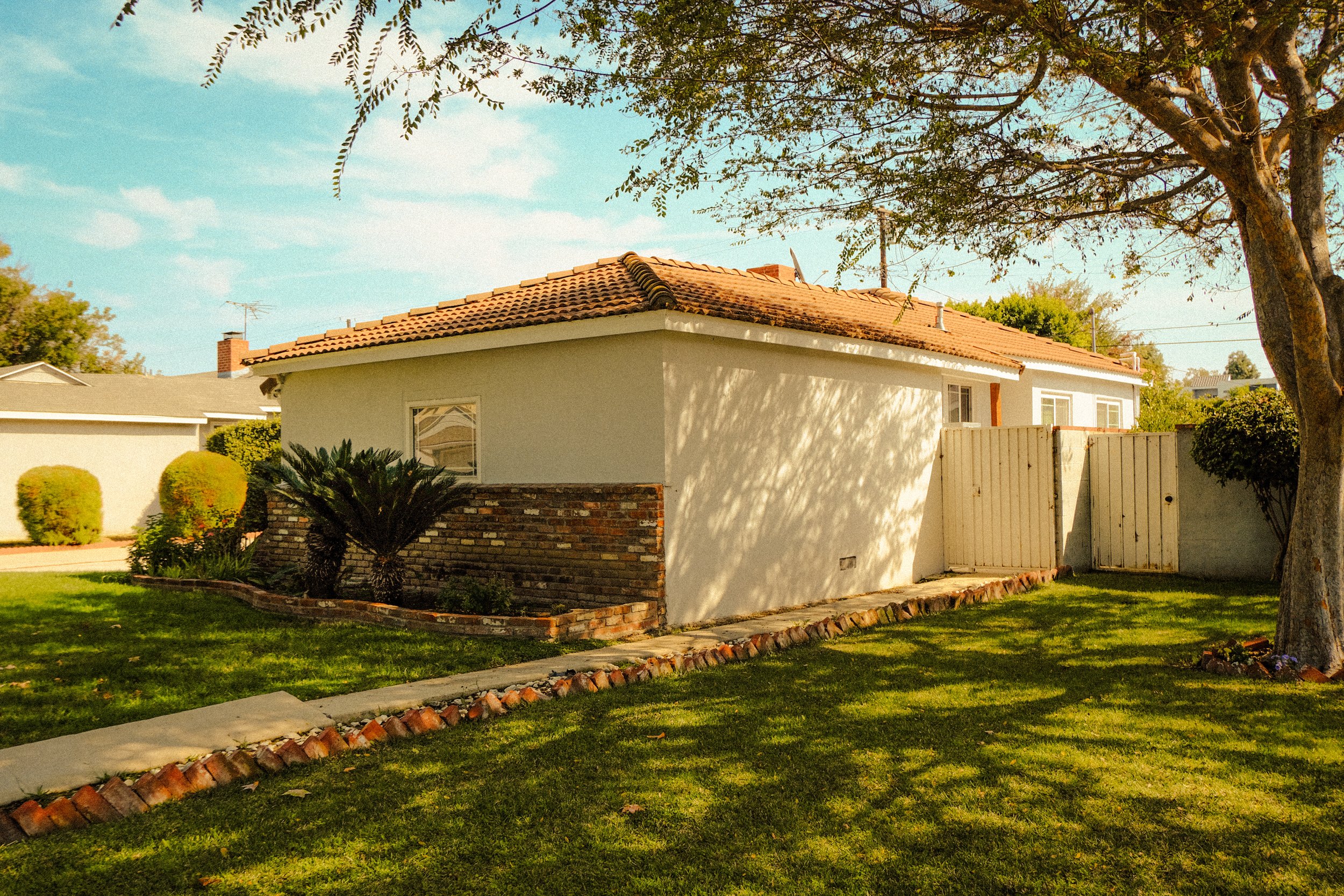 A suburban backyard with a single-story house, green lawn, garden bed with small palm trees, large tree providing shade, white gate, and blue sky with a few clouds.