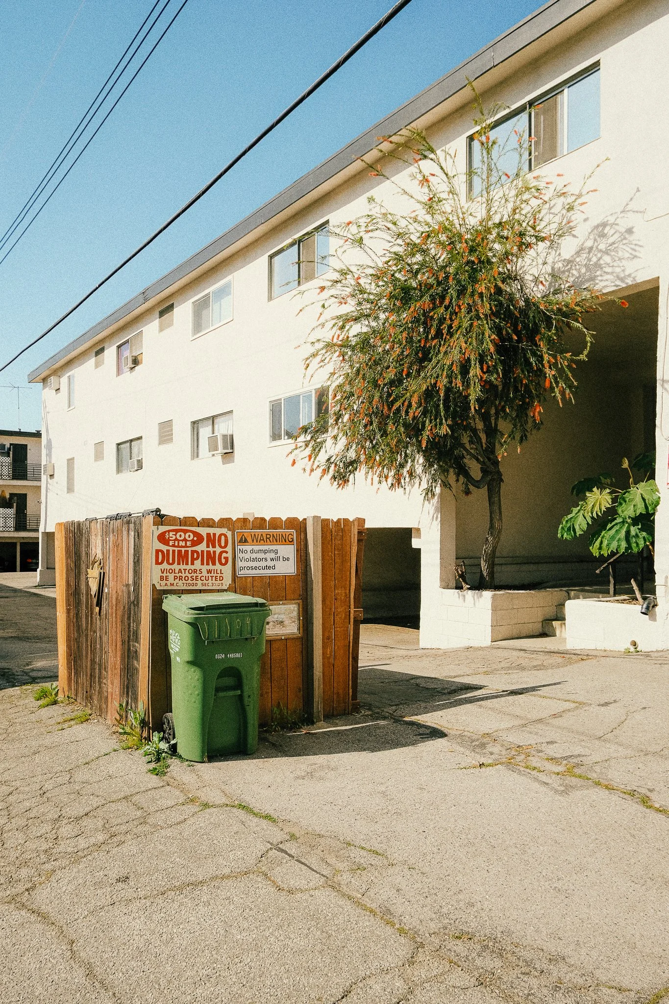 A white apartment building with multiple windows, a tree, a green trash bin, and a wooden fence with warning signs.