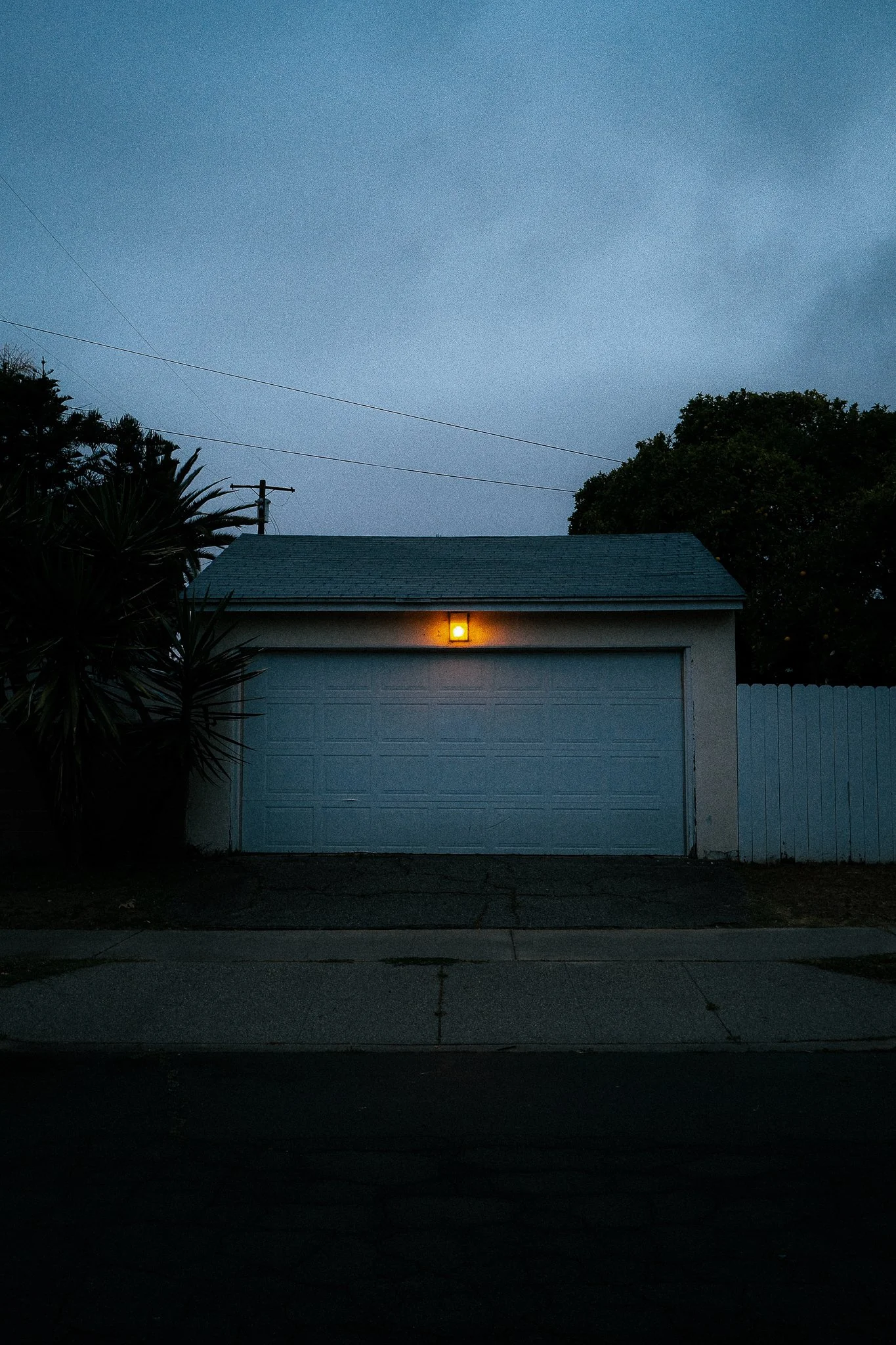 A garage with a closed white door, a small yellow light above it, surrounded by trees and a white fence, under a dark, cloudy sky.
