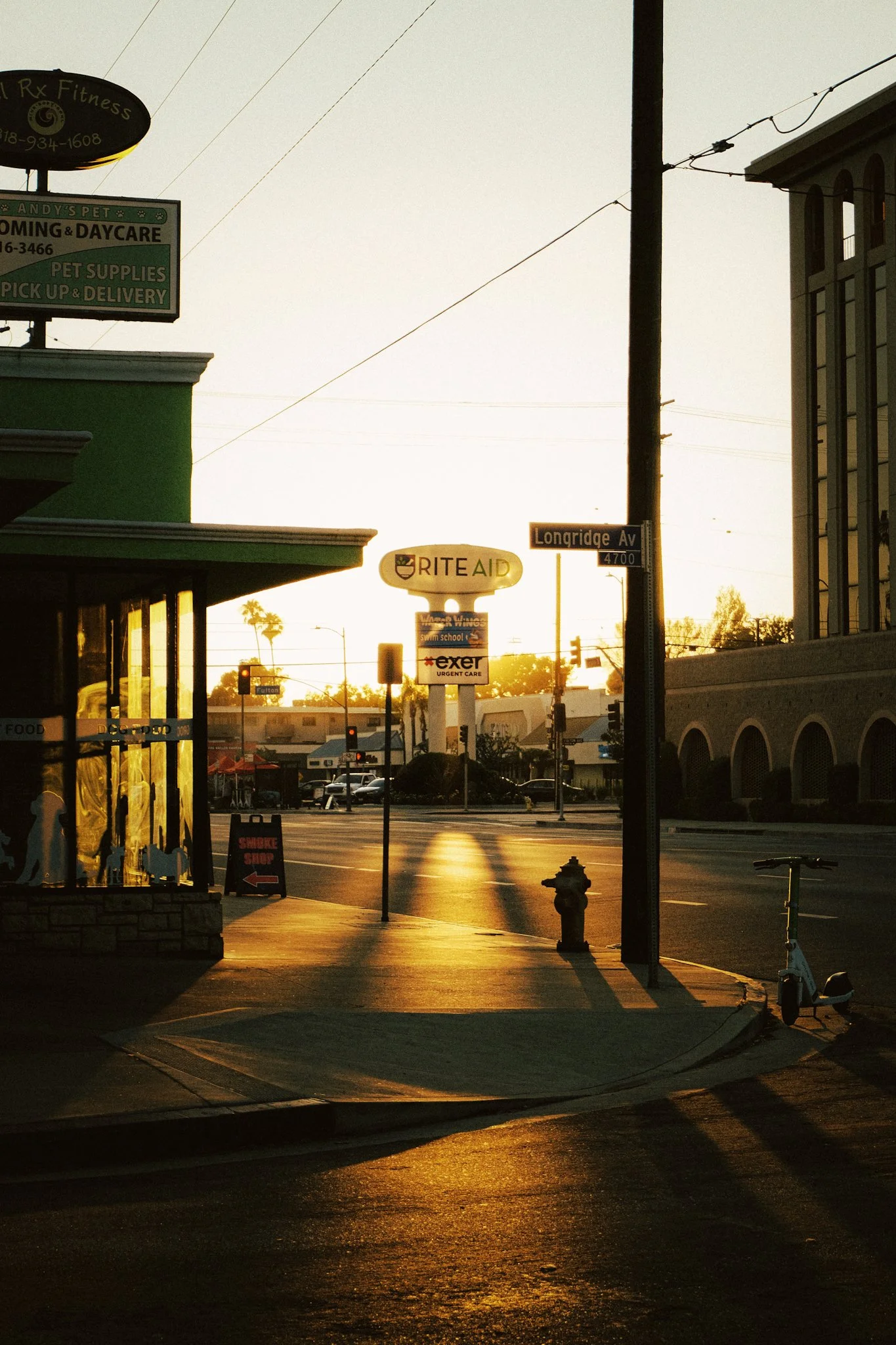 Sunset view of a city street corner with storefronts, signs, and a parked scooter, bathed in warm golden light.