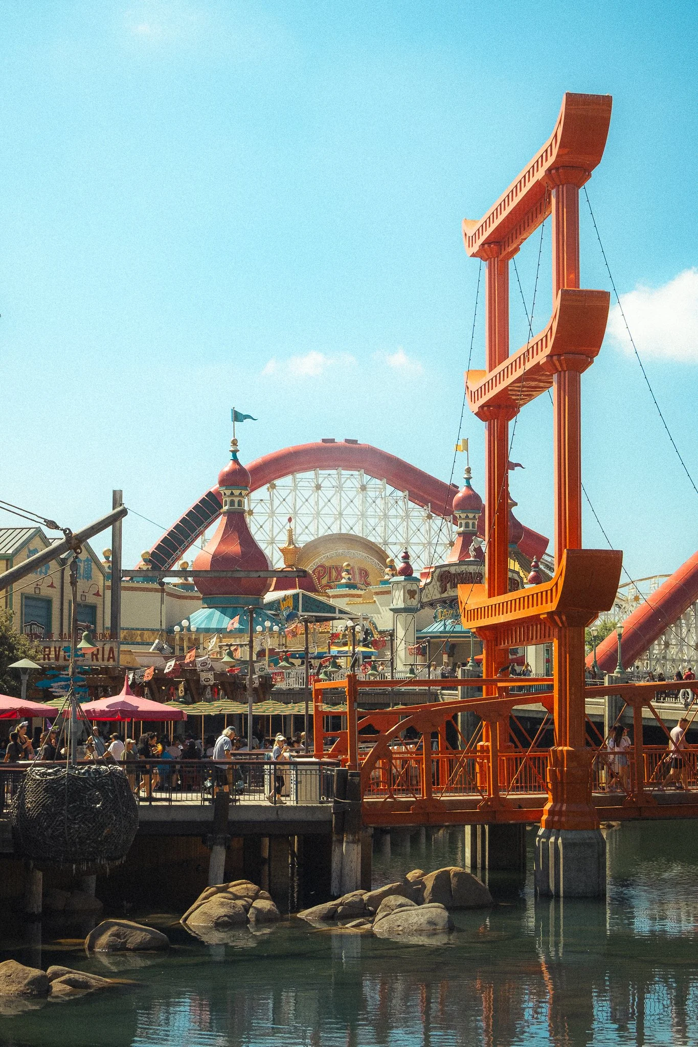 View of an amusement park with a prominent orange gondola ride structure in the foreground, a crowd of people walking on the pier, and a large roller coaster in the background under a clear, blue sky.