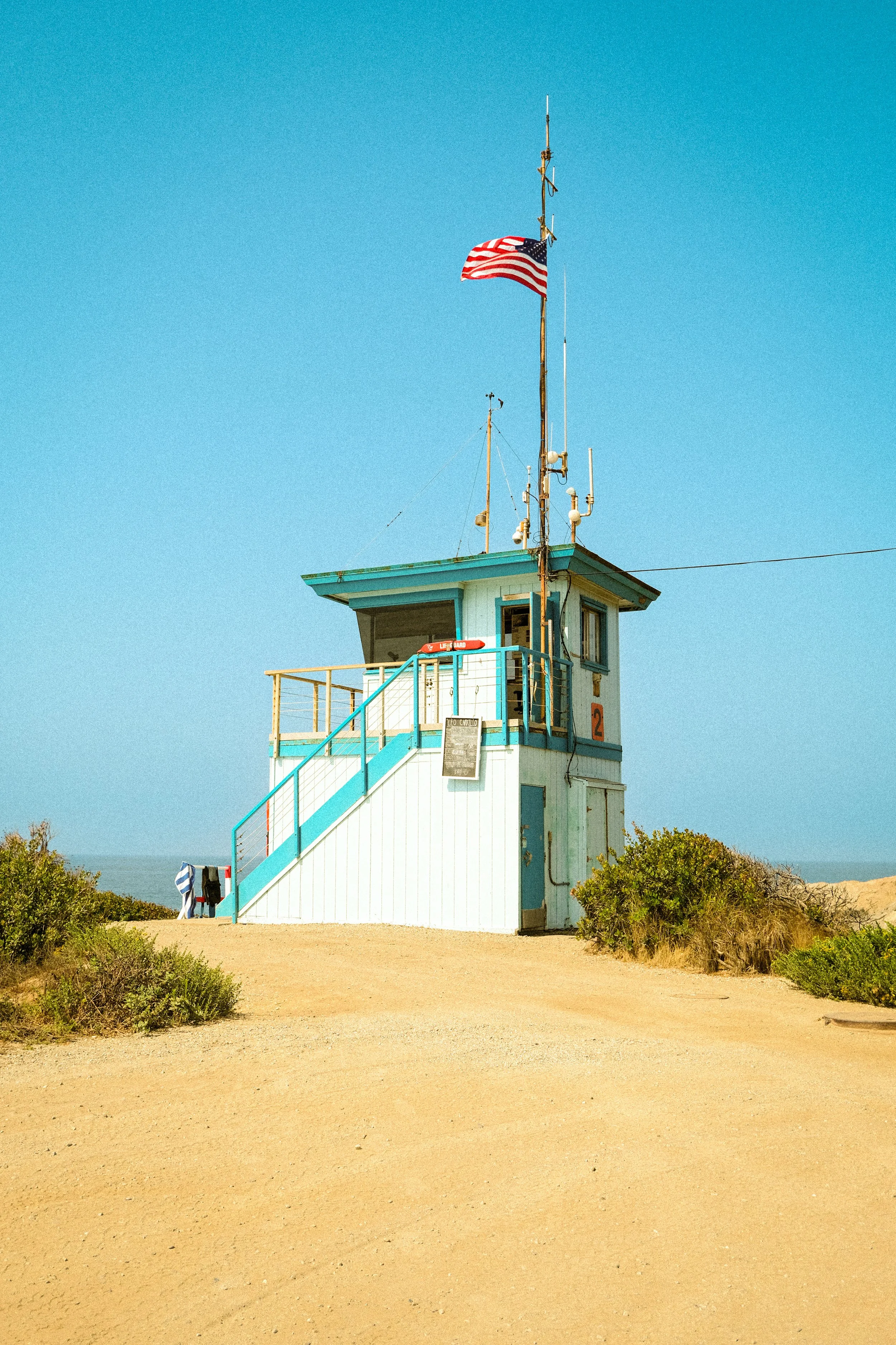 A small, light blue and white lifeguard tower on a sandy beach, with an American flag flying atop a tall pole against a clear blue sky.