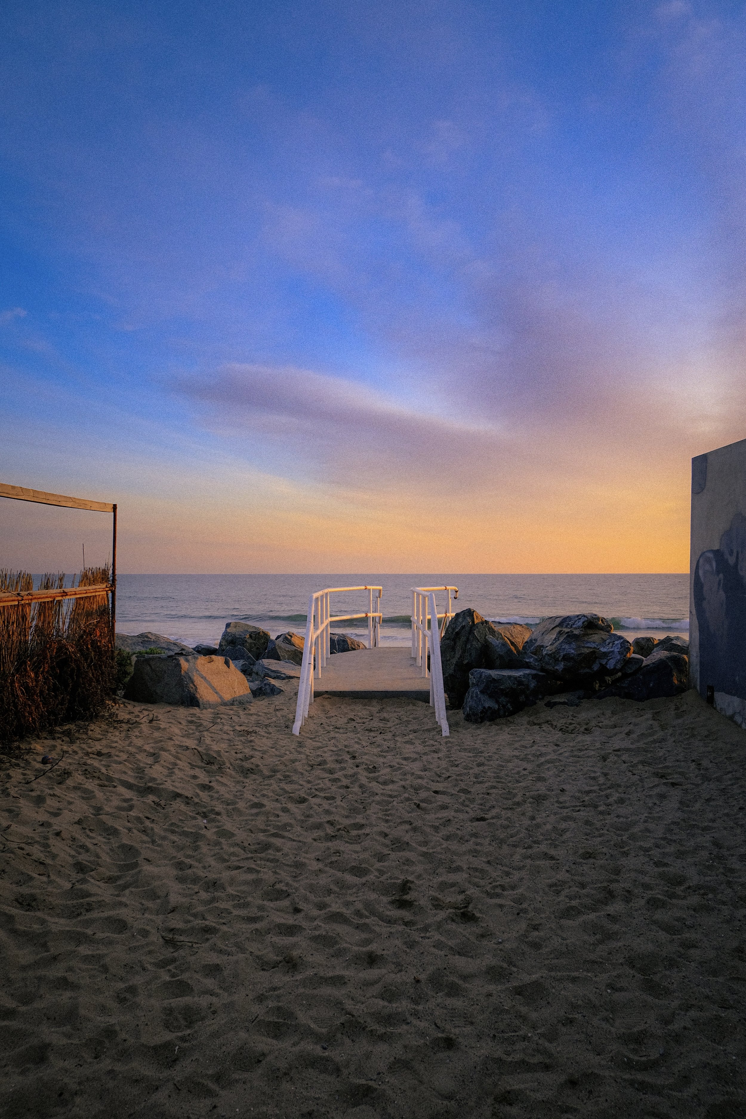 A sandy beach with a small wooden pier leading to the ocean, rocks on either side, and a sunset sky with blue and orange hues.