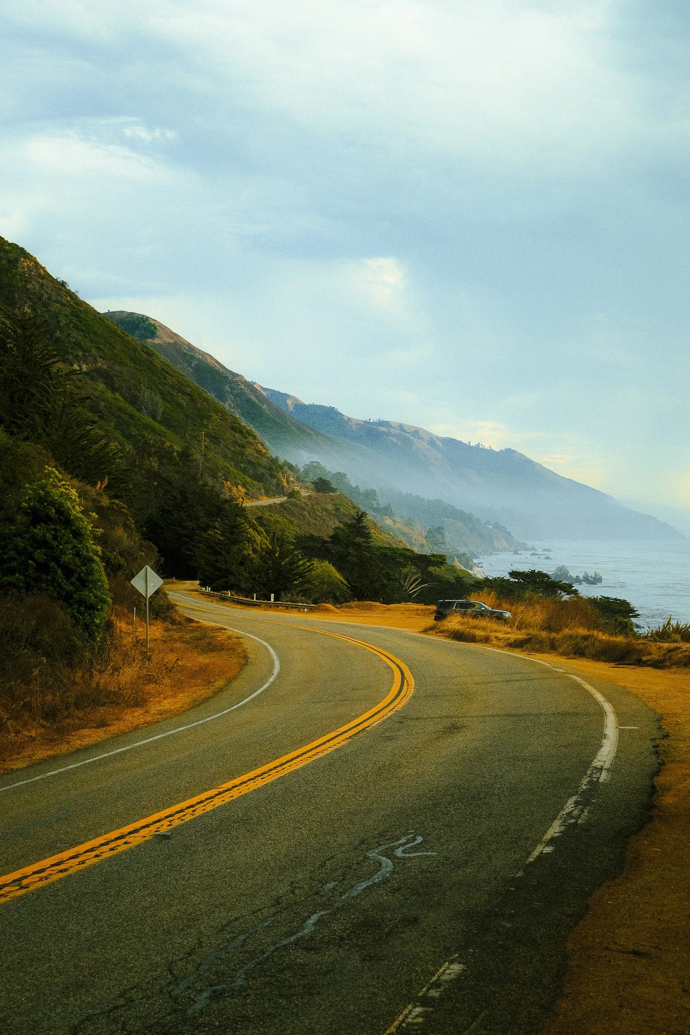 A winding coastal road running along a hillside with green trees,-facing the ocean with cliffs in the distance, under a cloudy sky.