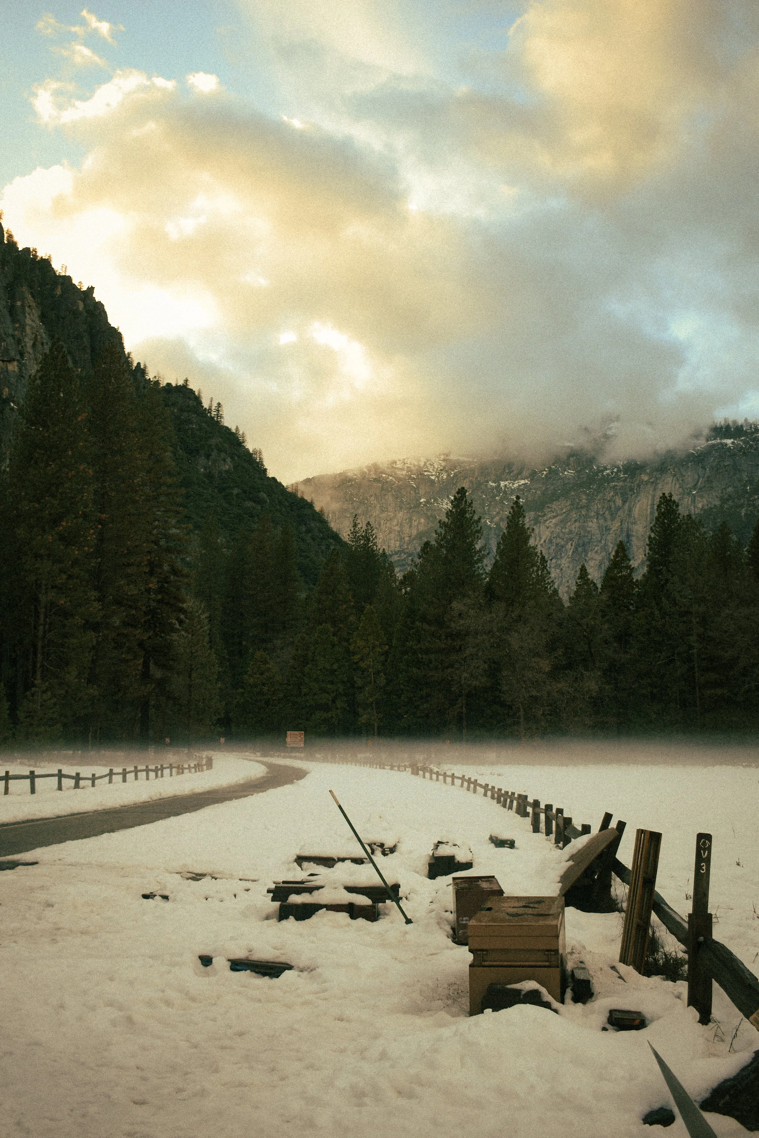 Snow-covered roadside in a forested mountain area with a guardrail, a broken street sign, and scattered debris on the snow, with a misty mountain and cloudy sky in the background.