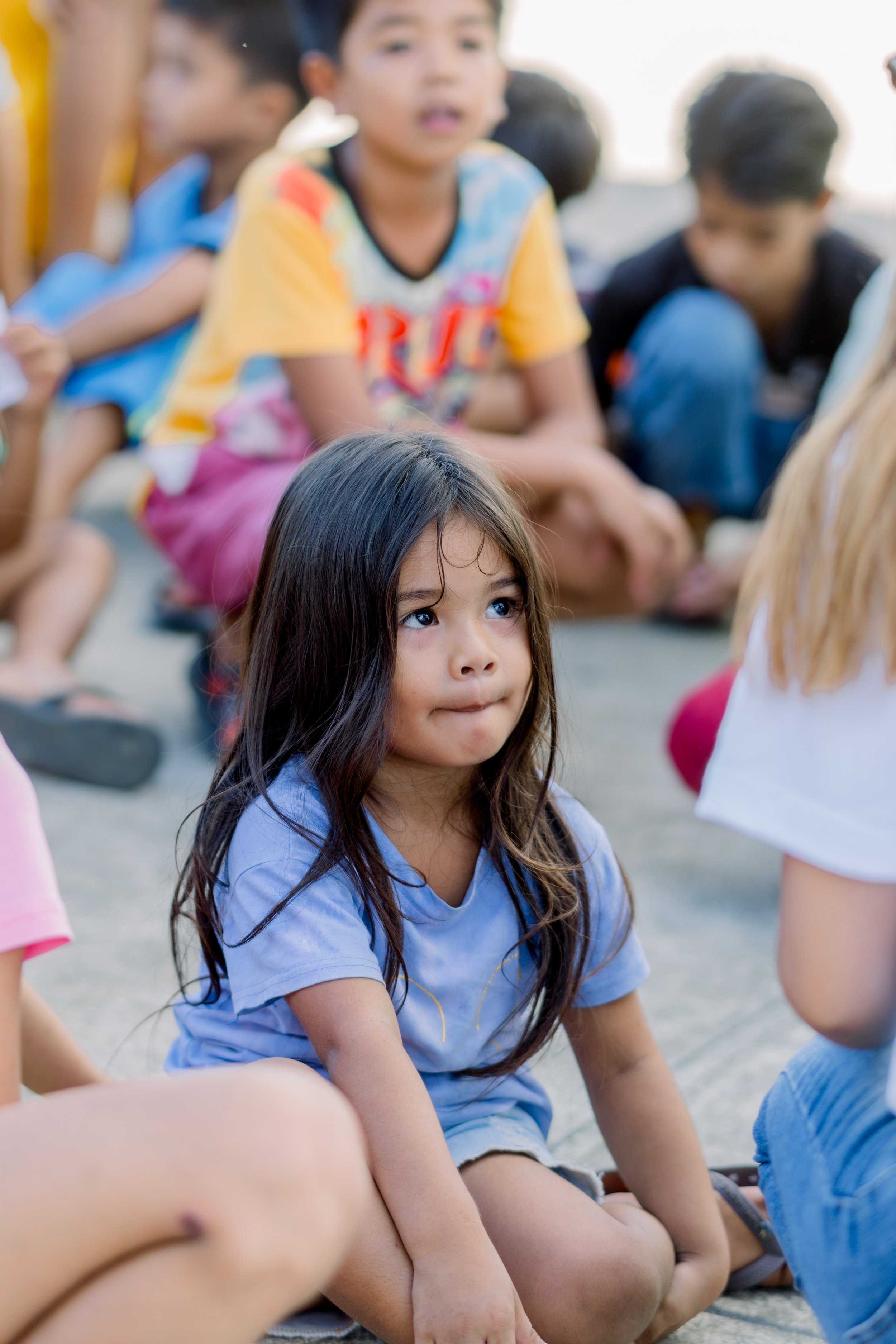 A young girl with long dark hair sitting on the ground, looking thoughtful and focused. She is wearing a light blue shirt, surrounded by other children sitting nearby.