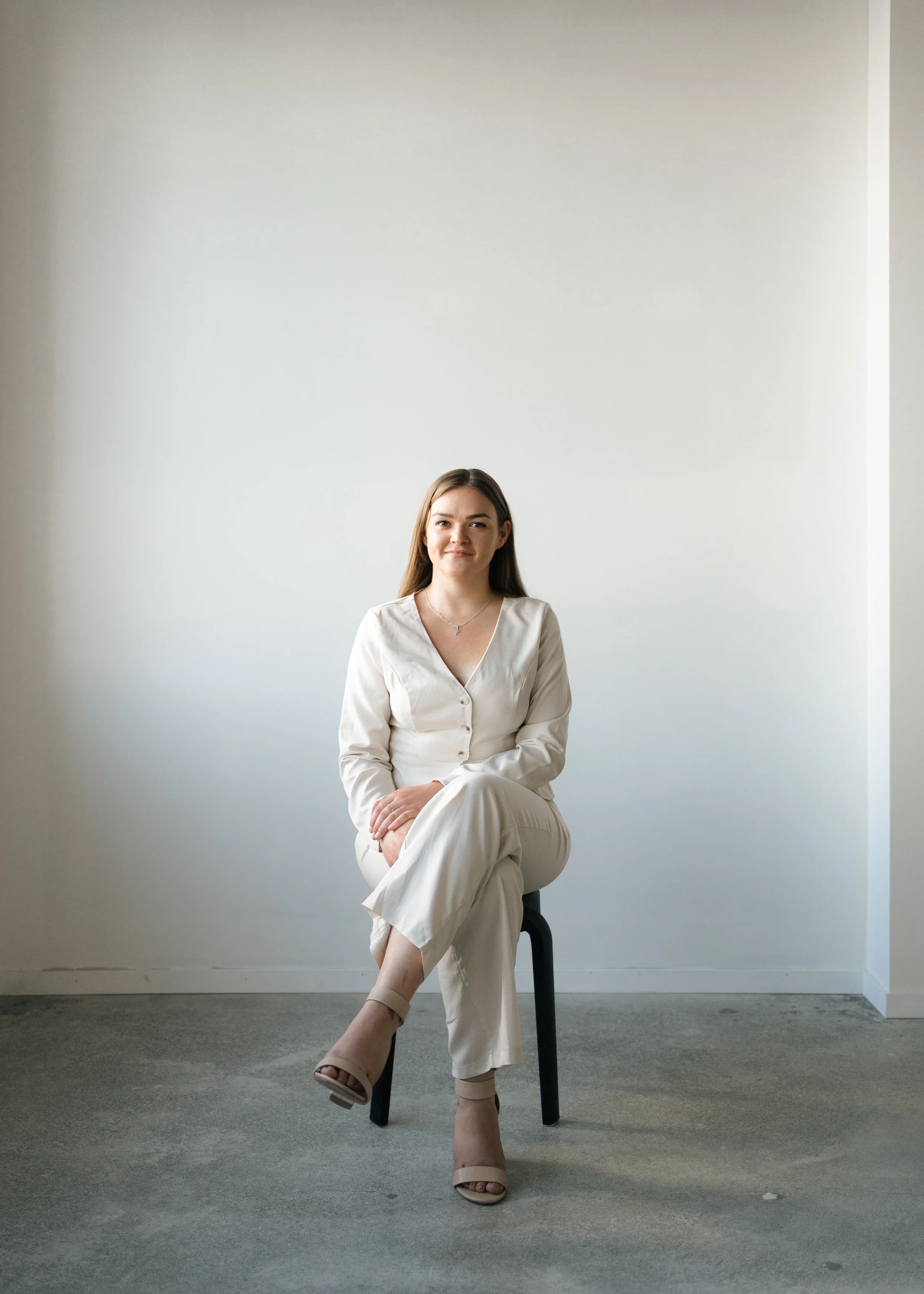 A young man with a mustache and beard, wearing a backward cap, a beige jacket, and teal pants, stands on a rooftop holding an acoustic guitar across his chest. The background features a cityscape under a cloudy sky during sunset.