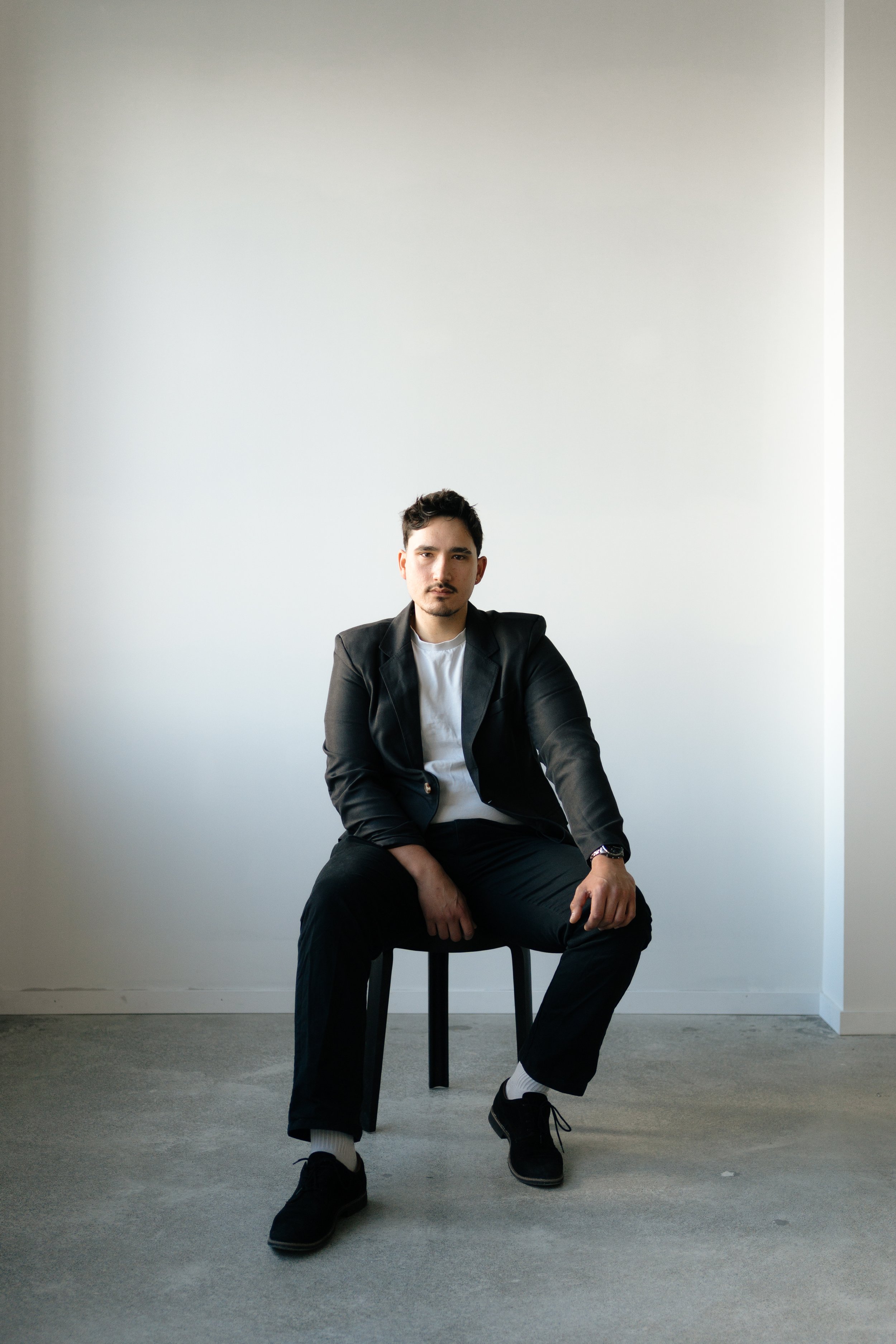 A young man standing outdoors on a balcony or rooftop, holding an acoustic guitar, with a cityscape in the background during sunset or late afternoon.