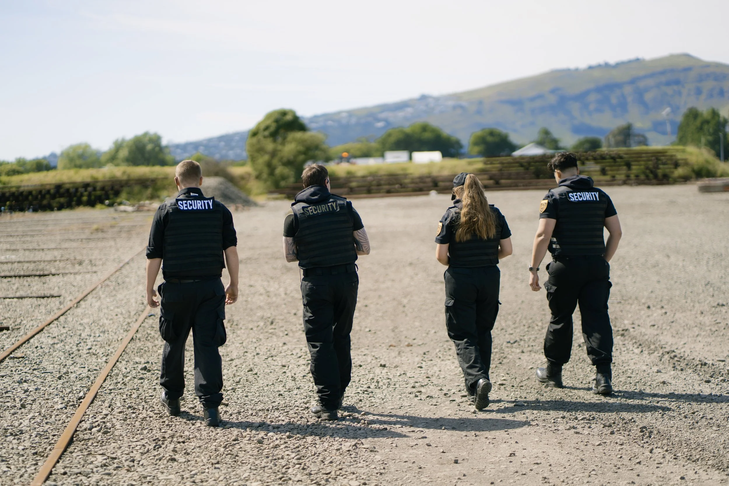 Four security guards walking along train tracks in an outdoor area with a mountain in the background.