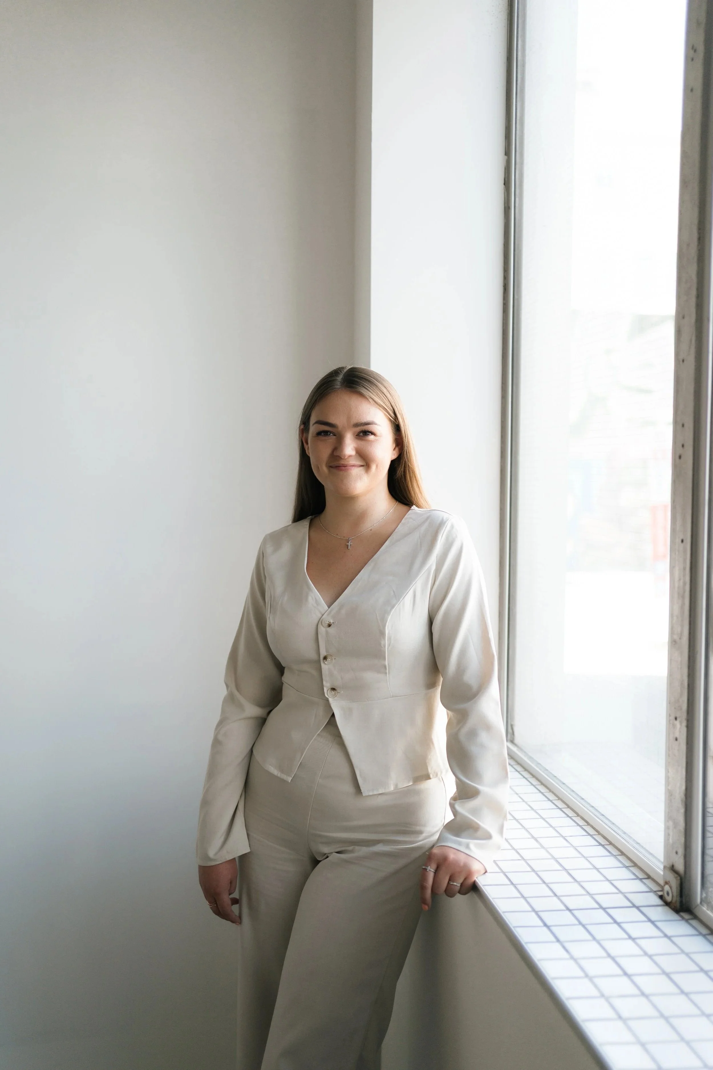 A young woman with long brown hair standing by a large window in a white room, wearing a matching beige suit and smiling.