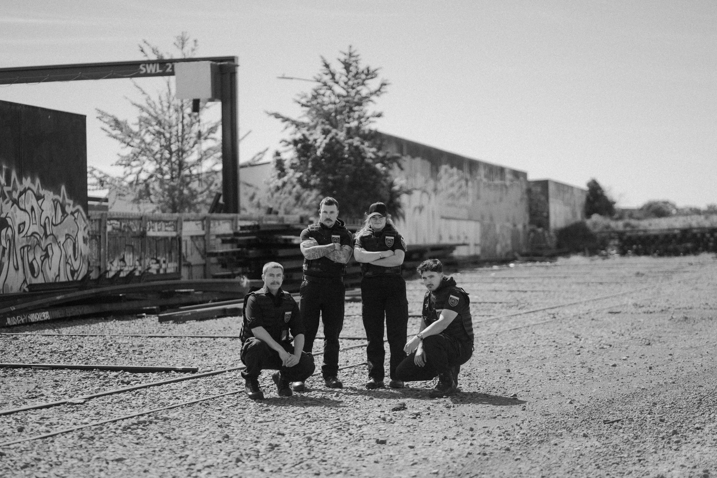 Four security officers in uniform, two kneeling and two standing with arms crossed, on a gravel surface in an urban outdoor area with graffiti-covered walls and trees in the background.