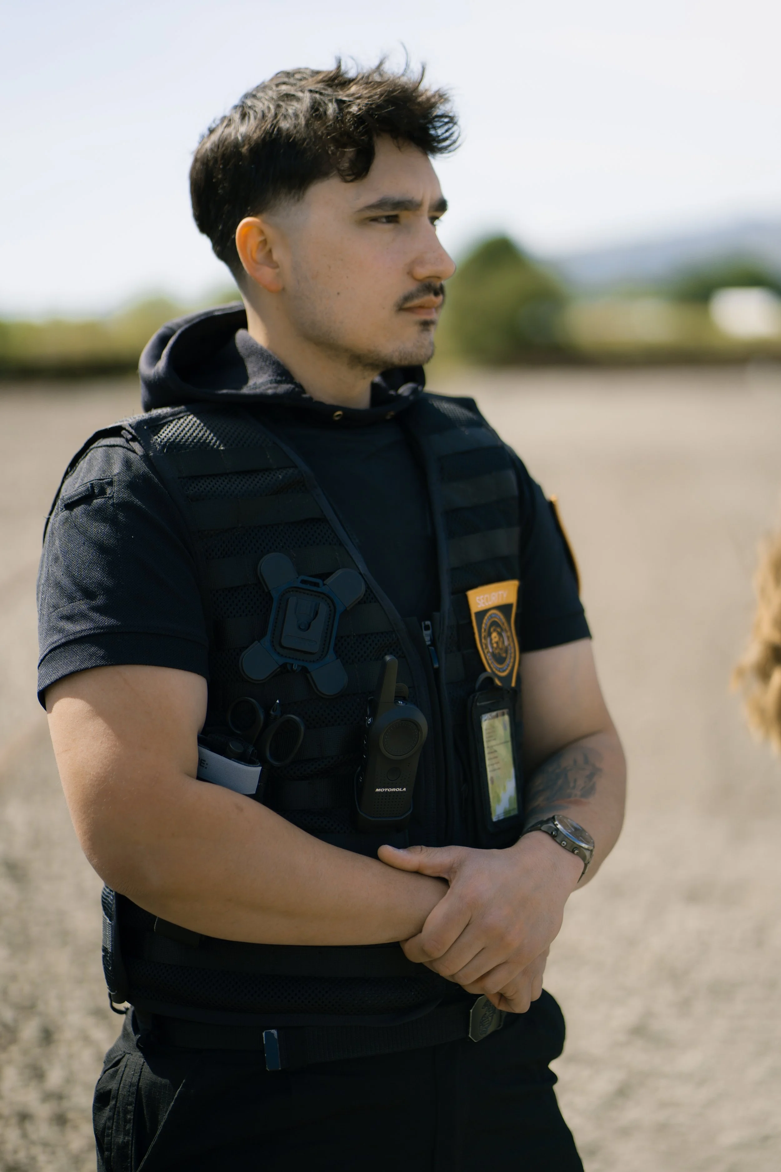 A male security officer standing outdoors during daytime, wearing a black uniform vest with security badges and equipment, including a radio and scissors, under a clear sky.