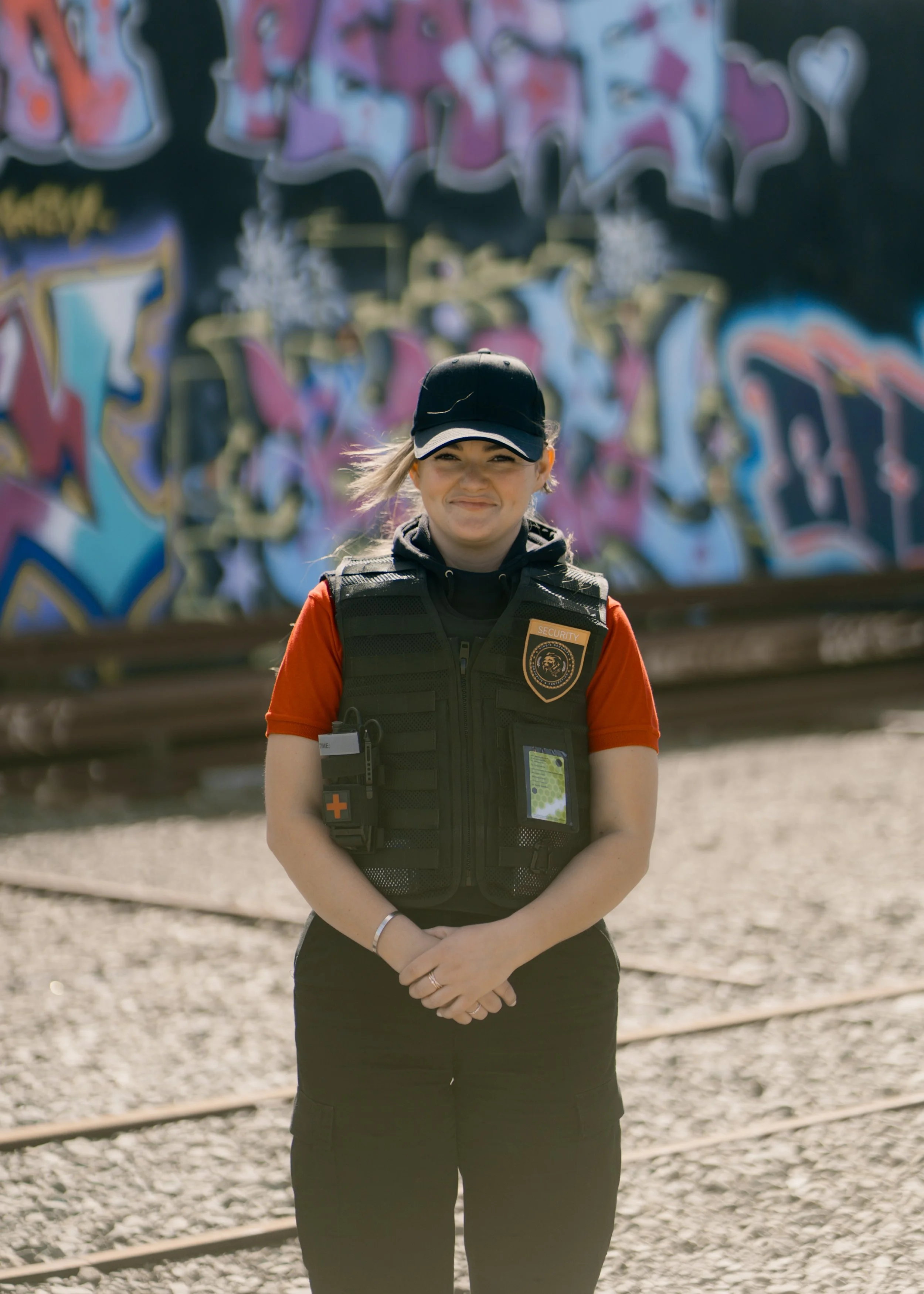 Security personnel woman standing outdoors near graffiti wall, smiling, wearing black vest with badges and red shirt.