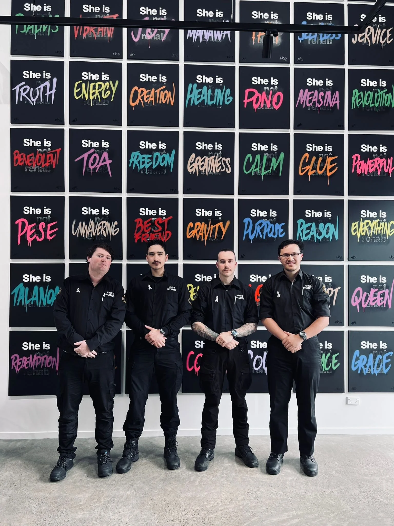 Four security personnel standing in front of a colorful wall display with motivational words, including 'Truth', 'Energy', 'Healing', 'Revolution', and 'Grace' among others, each word written in vibrant, graffiti-style text on black background panels.