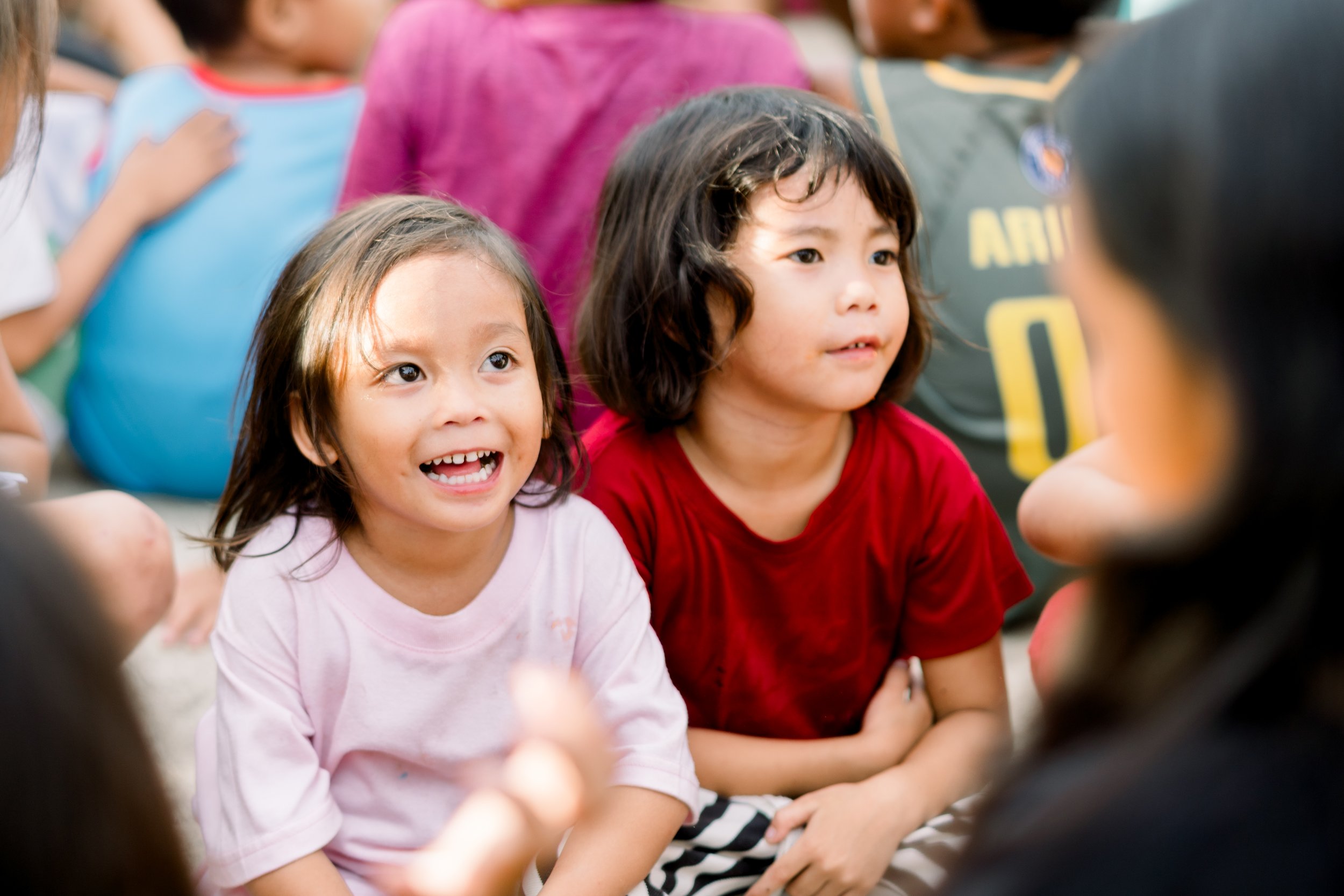 Two young girls sitting on the ground, engaging in conversation with a woman outside the camera frame, surrounded by other children.