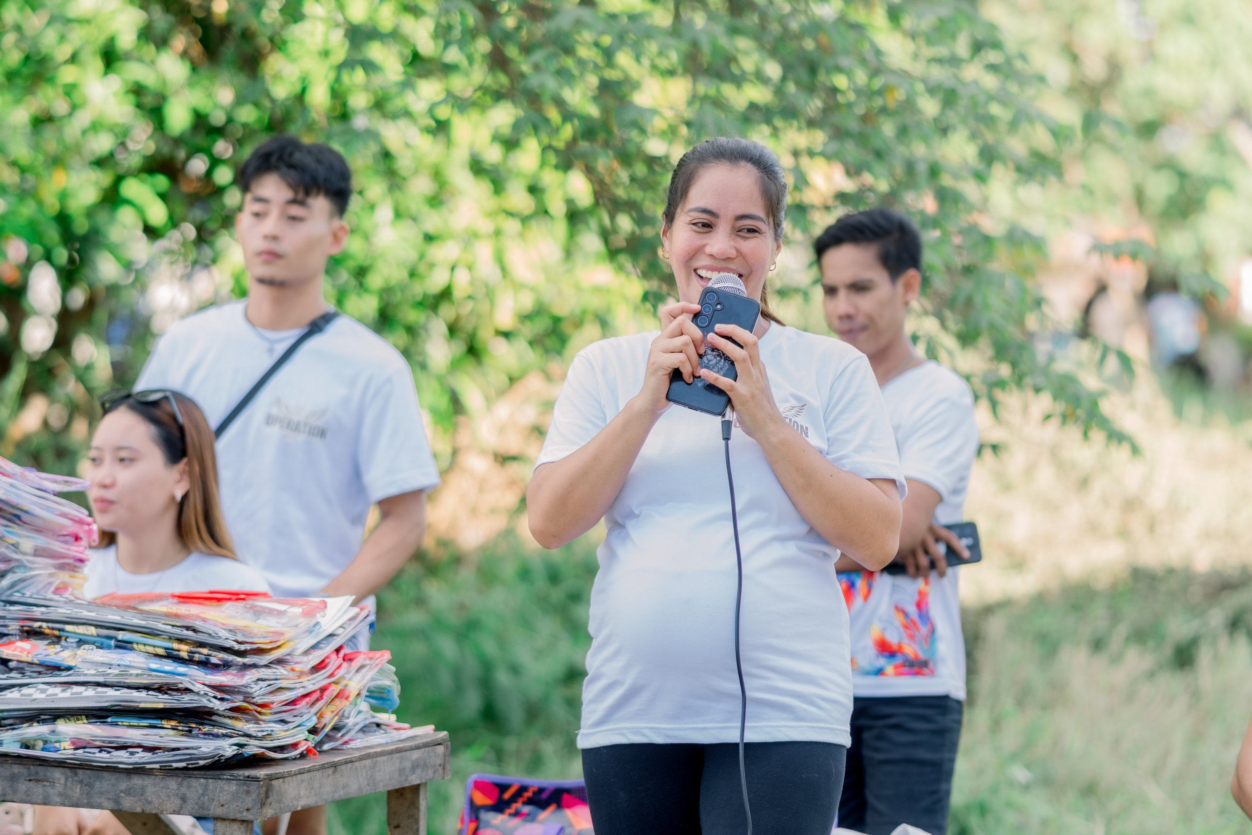 Smiling woman holding microphone and smartphone outdoors with group of people and greenery in background
