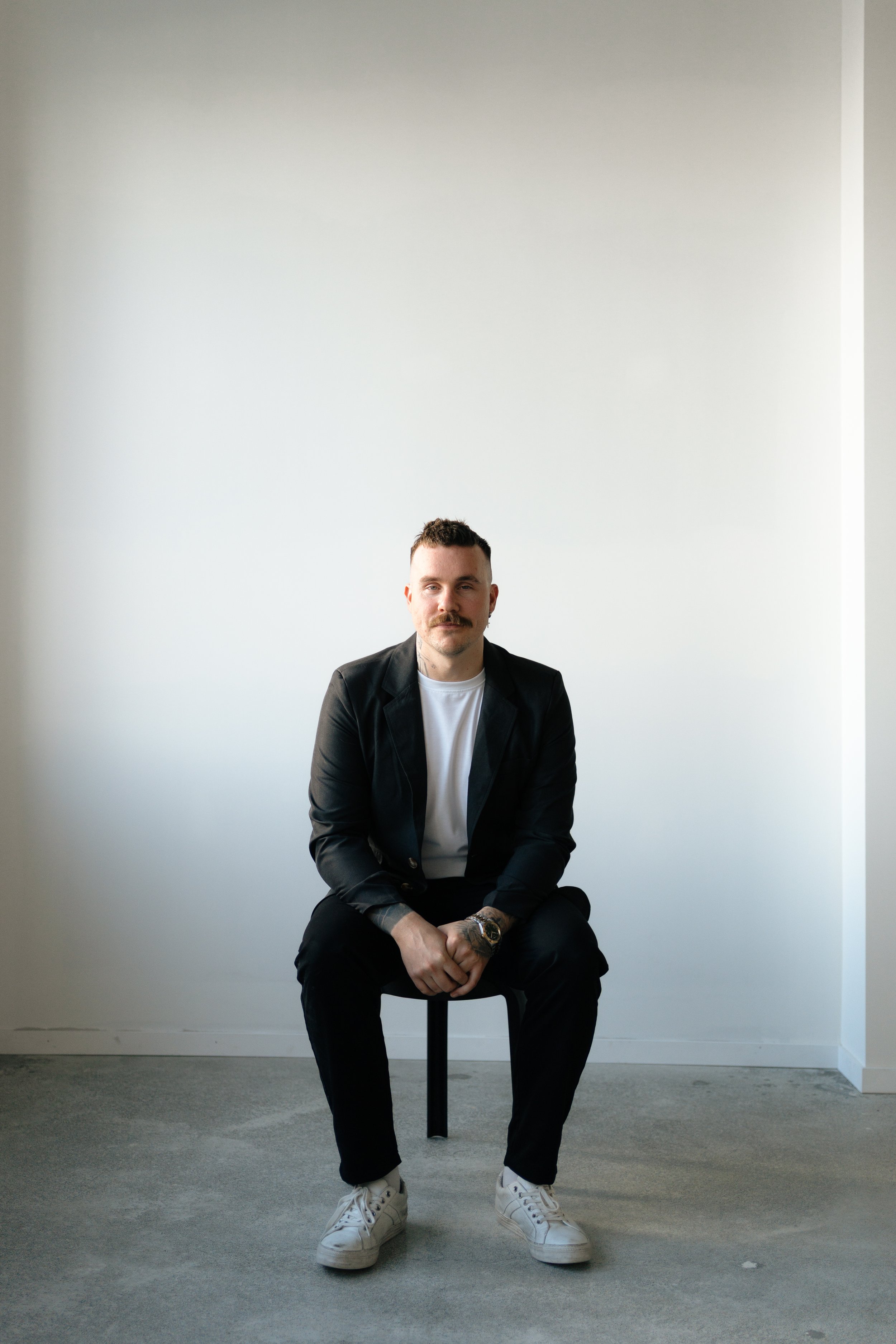A man with a mustache and beard wearing a black baseball cap backwards, plaid jacket, and dark pants, is holding an acoustic guitar on his shoulder while standing on a rooftop with city buildings in the background during sunset.