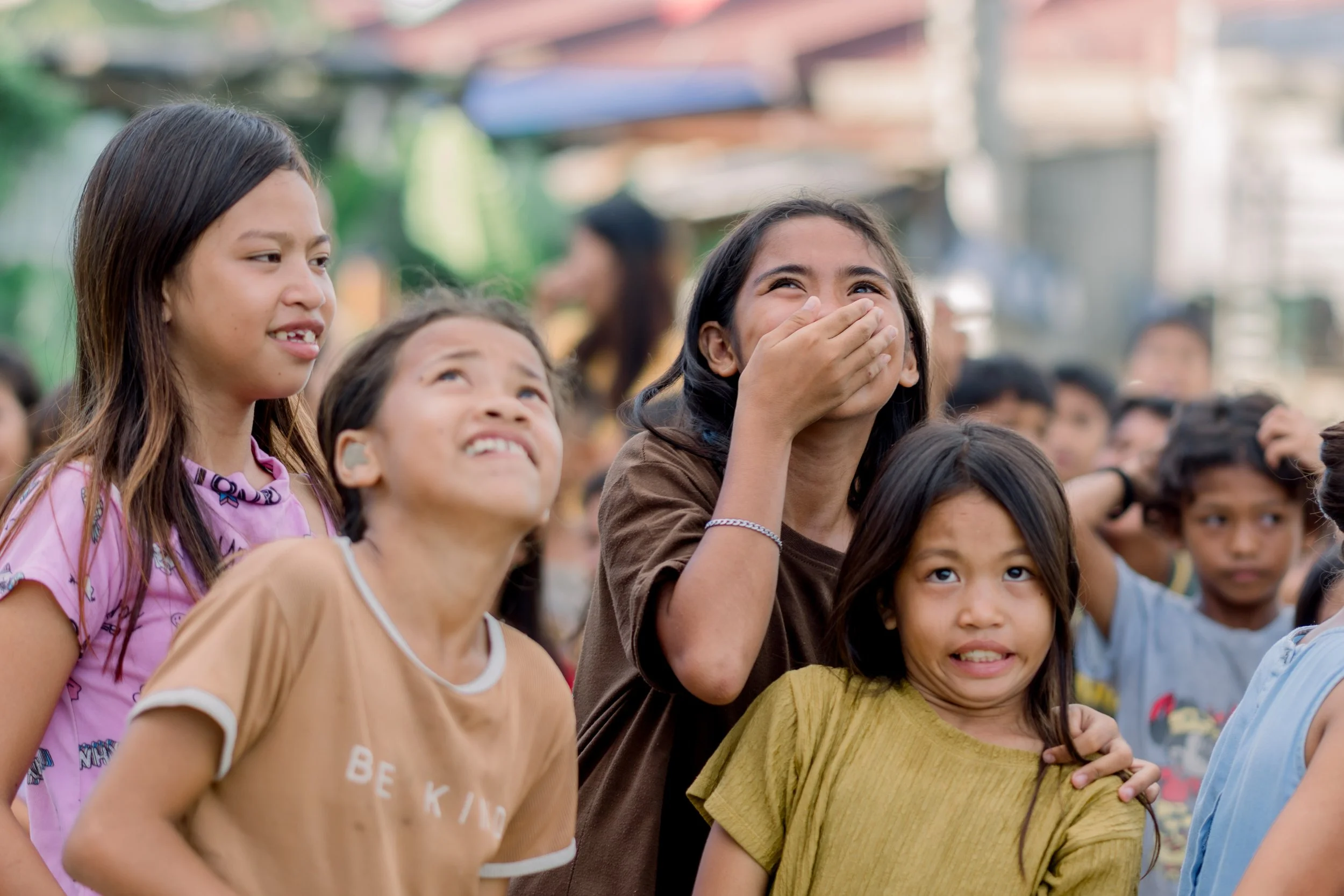 Group of children outdoors, some laughing and smiling, with a blurry background of a crowd and buildings.