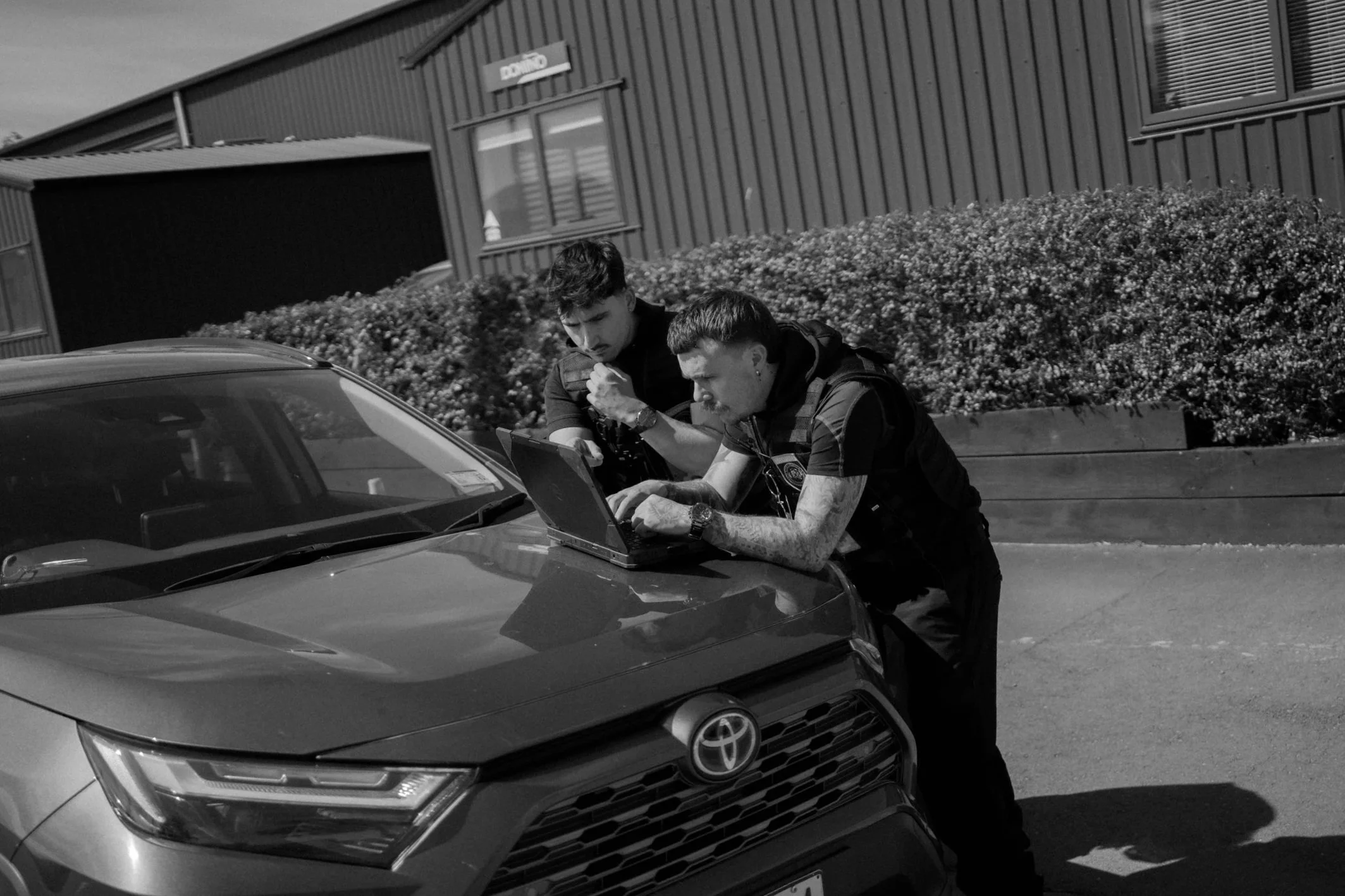 Two security officers studying a laptop on the hood of a Toyota car outside a building with bushes in the background.