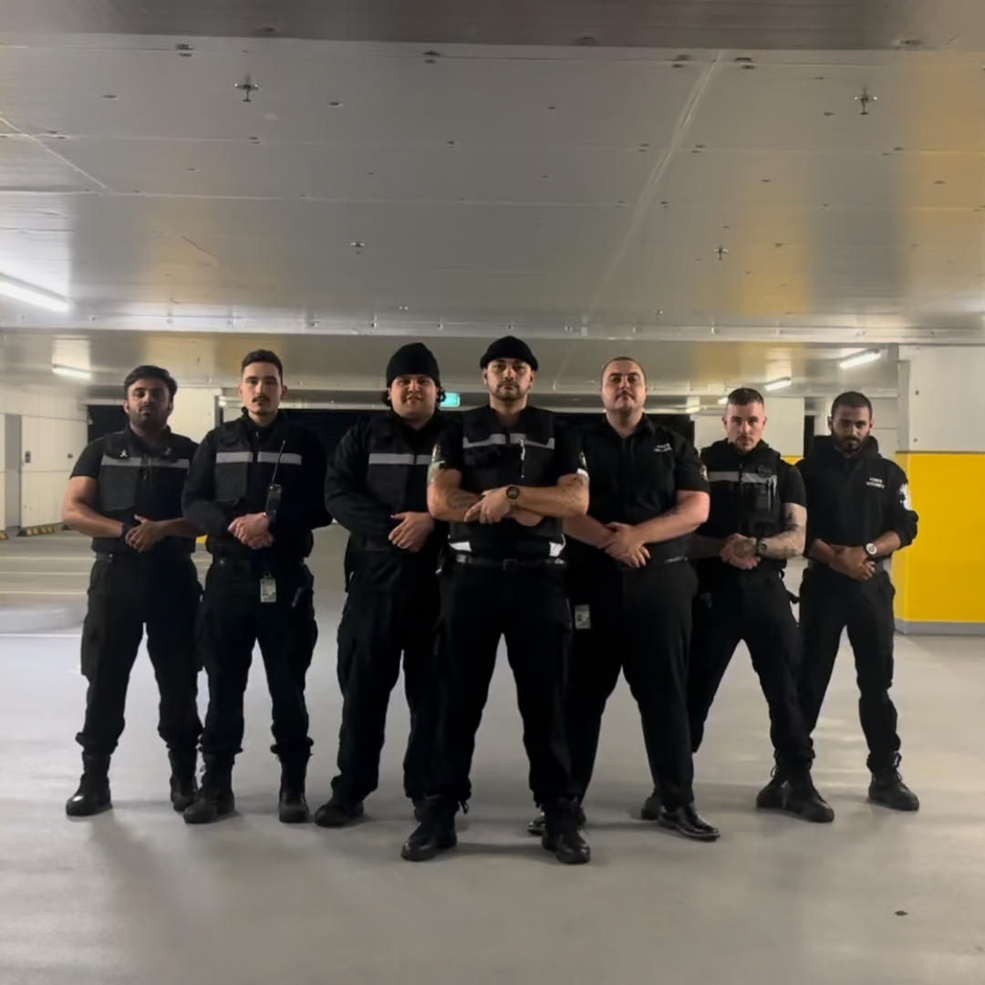 Group of seven security guards standing together in an underground parking garage, dressed in black uniforms with reflective stripes, with serious expressions and arms crossed or at their sides.