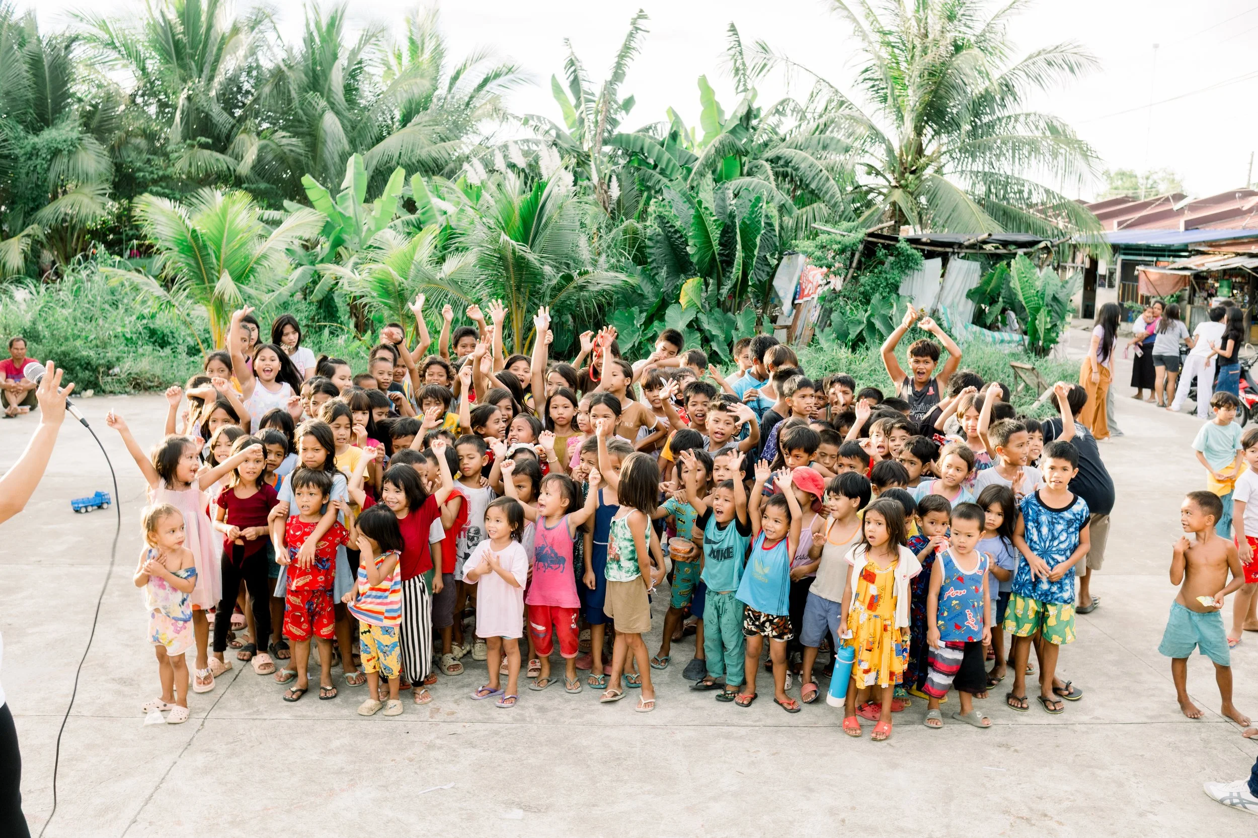 A large crowd of children and some adults gathered outdoors on a concrete area with lush green tropical plants in the background. The children are smiling, raising their hands, and appear to be participating in an event or activity.