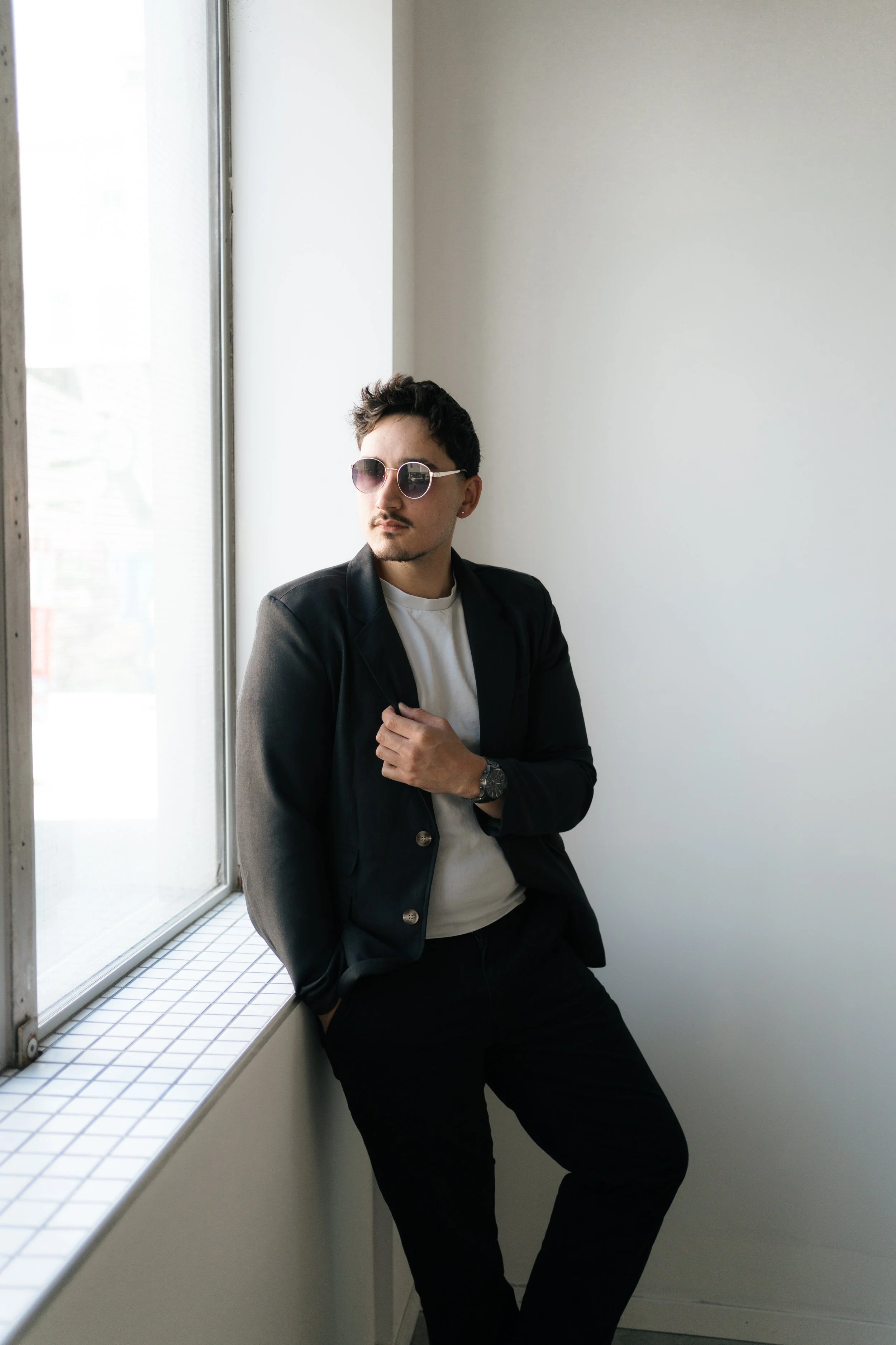 A man leaning against a windowsill in a minimalist room, wearing sunglasses, a black blazer, white t-shirt, black pants, and a watch.