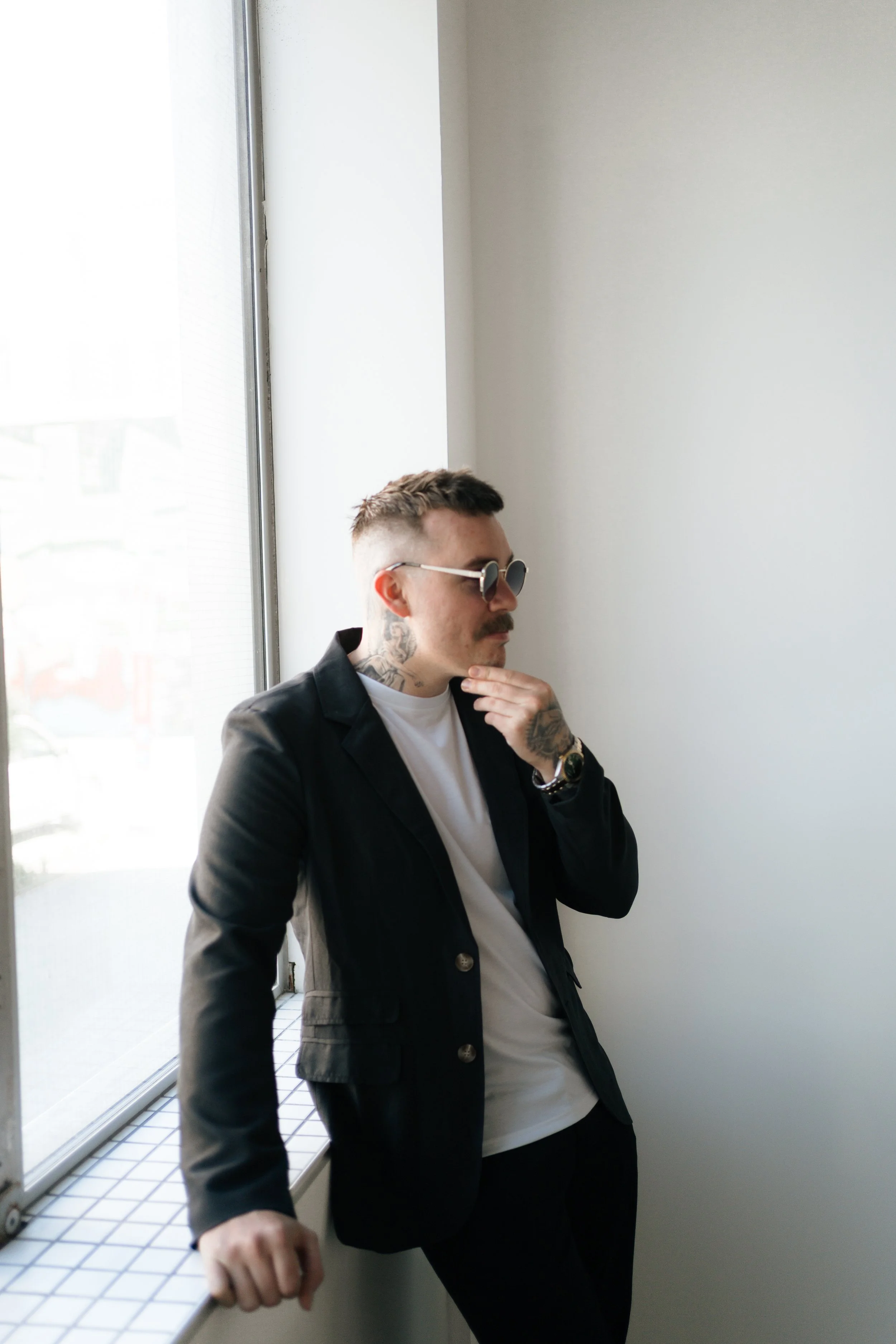 A man wearing sunglasses, a white t-shirt, and a black blazer, leaning against a windowsill indoors, with tattoos visible on his neck and hand.