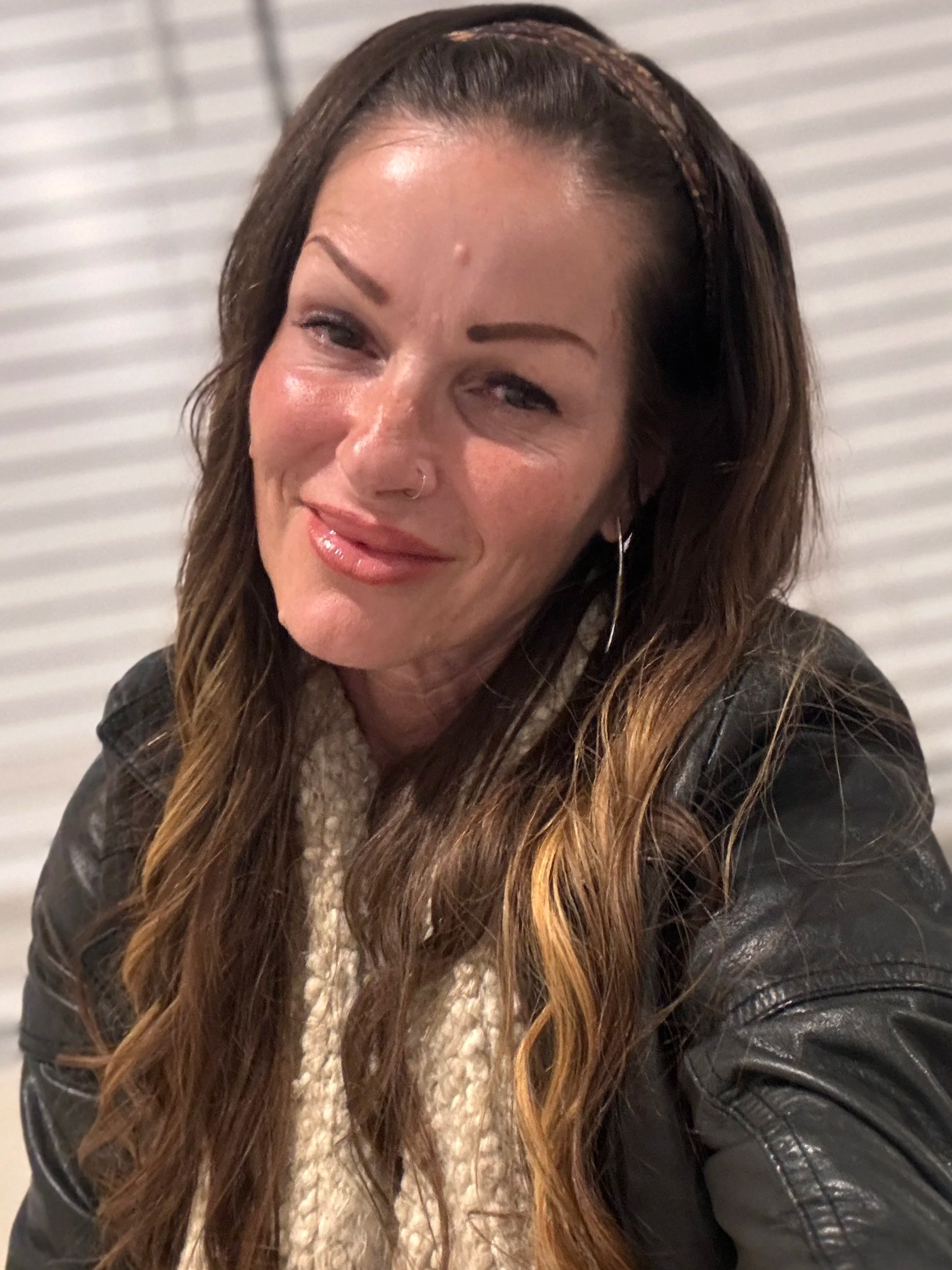 A woman with long brown hair wearing a black leather jacket, a beige scarf, earrings, and a headband.