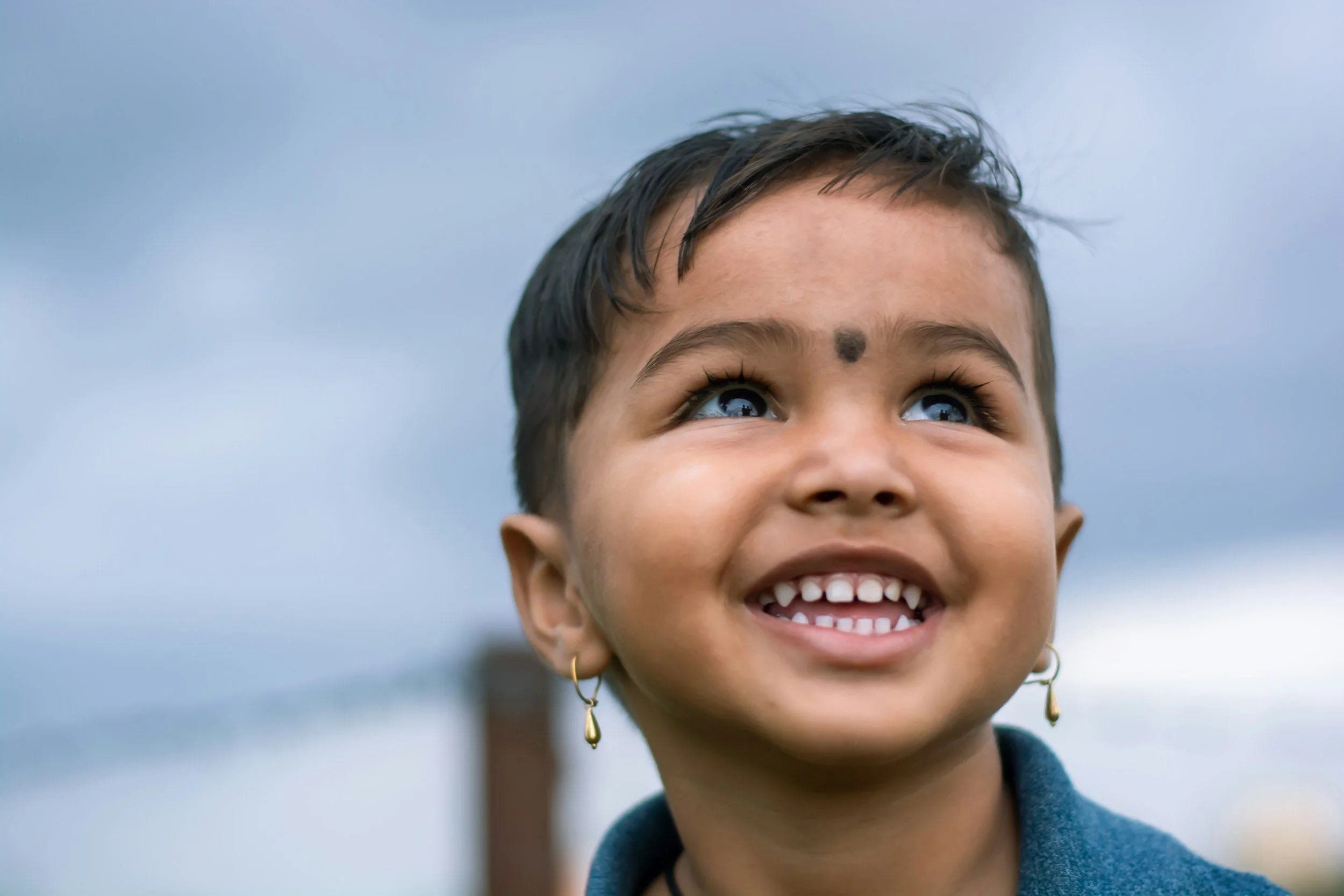 Close-up of a smiling young girl with short dark hair and blue eyes, wearing earrings and looking up, outdoors on a cloudy day.