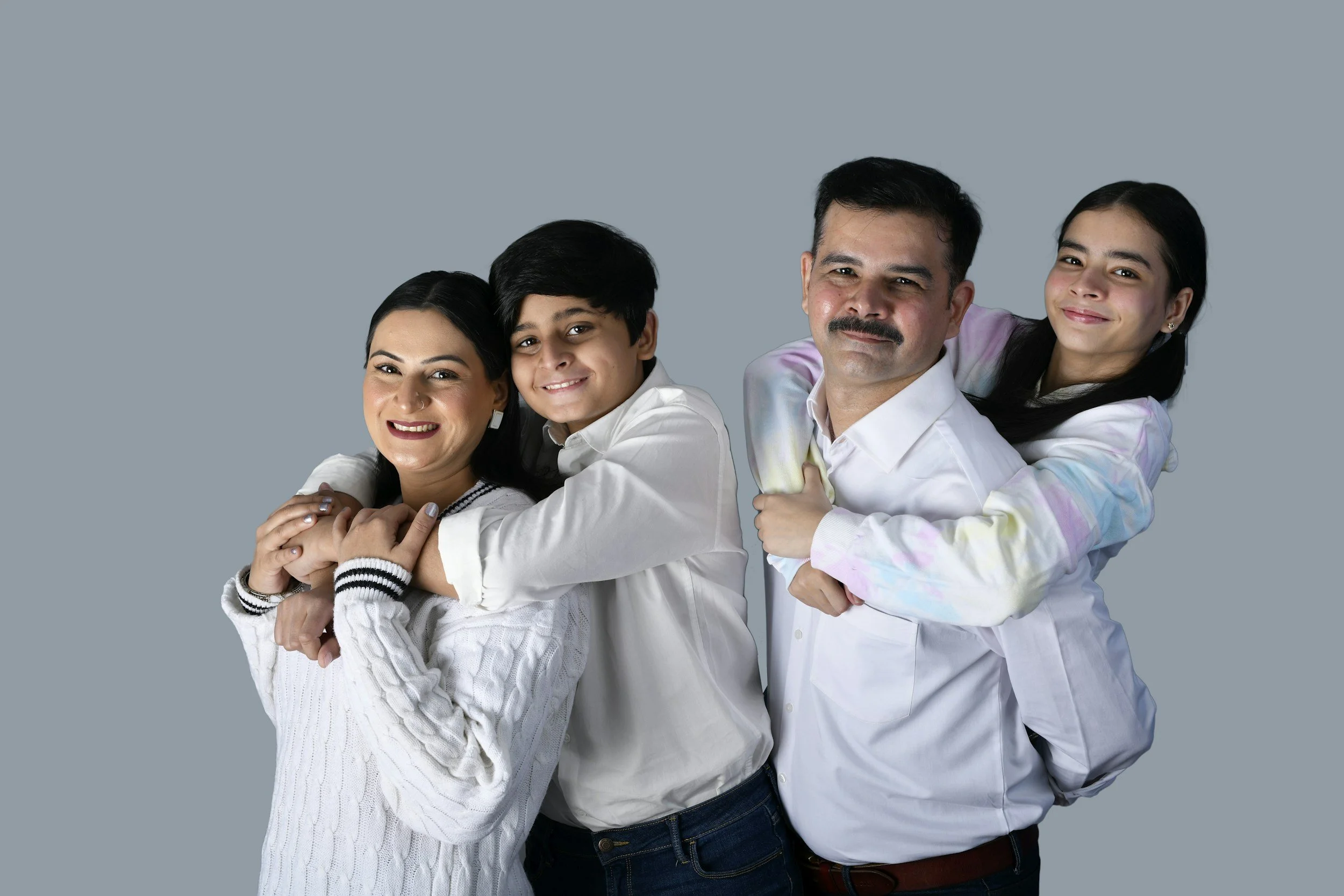 Family of four smiling, with two girls piggybacking on their parents, against a plain gray background.