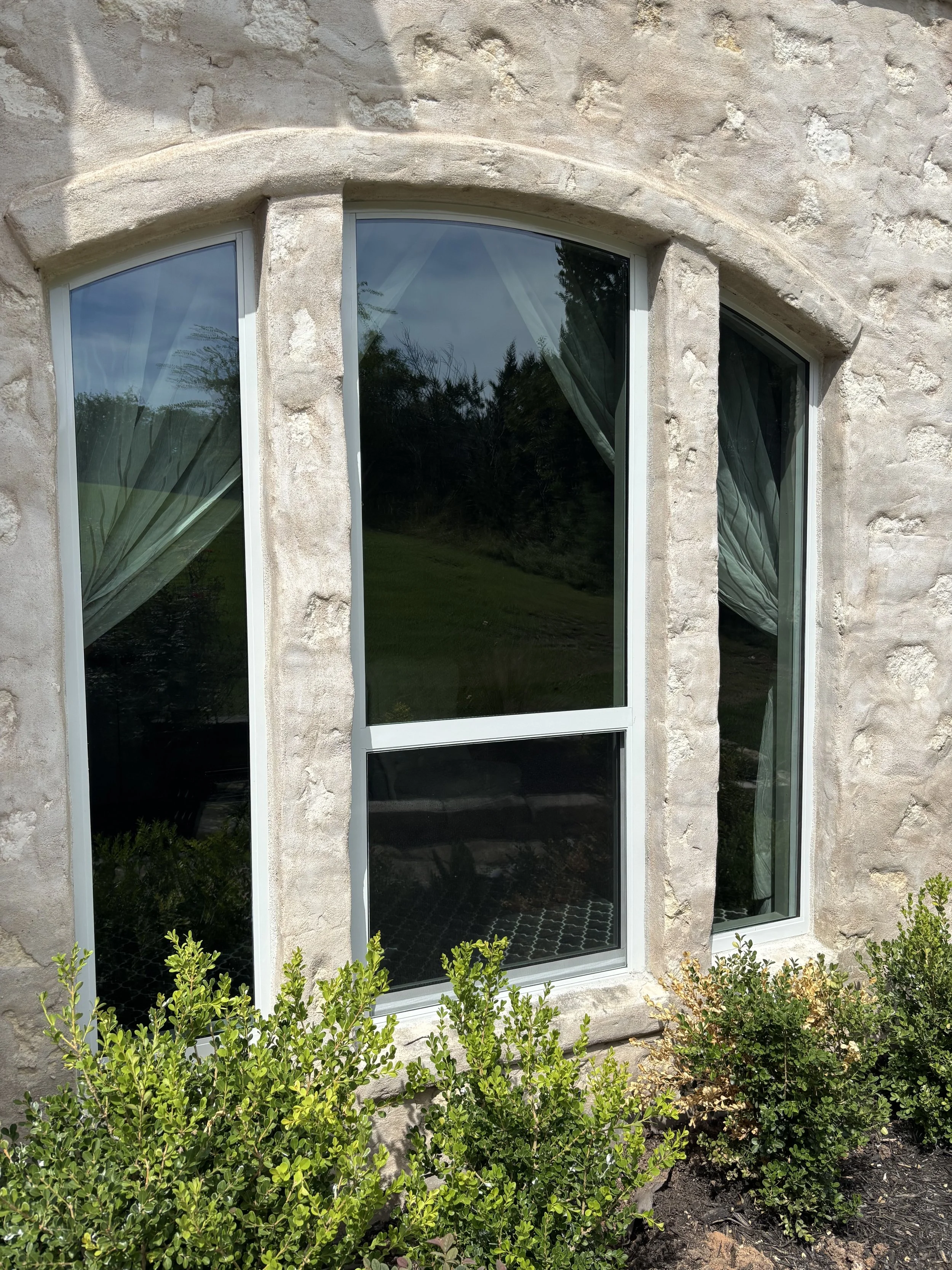 A close-up view of a stone house exterior featuring large, arched glass windows with white frames, with green shrubs in the foreground.