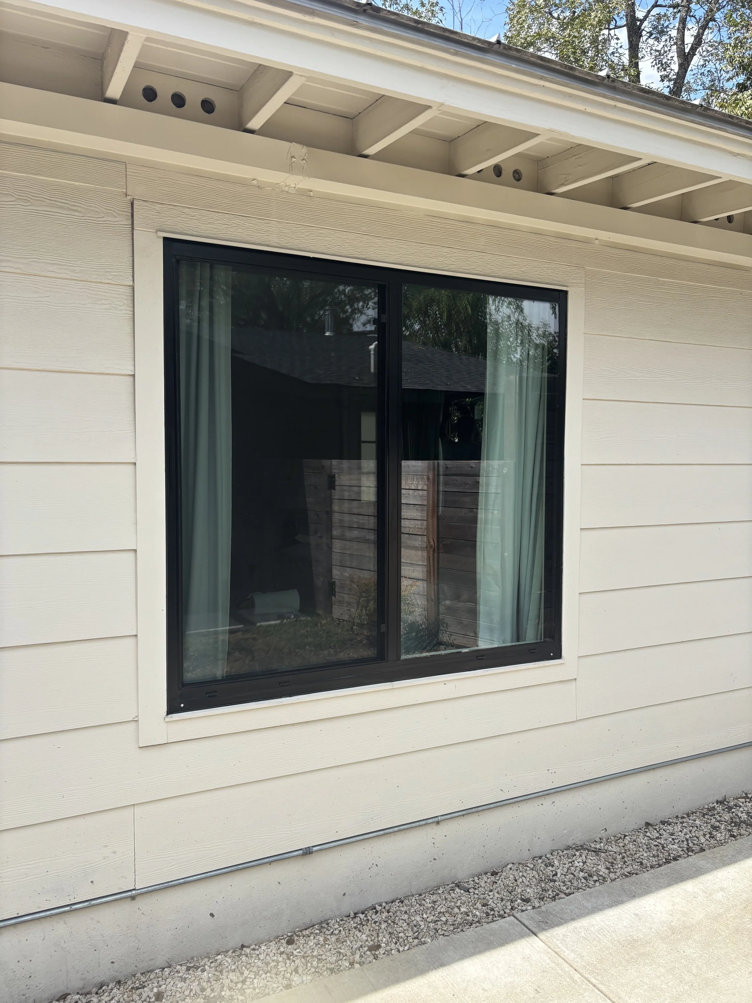 A house exterior with a large black-framed sliding glass window, beige siding, gravel along the foundation, and a concrete sidewalk in front.