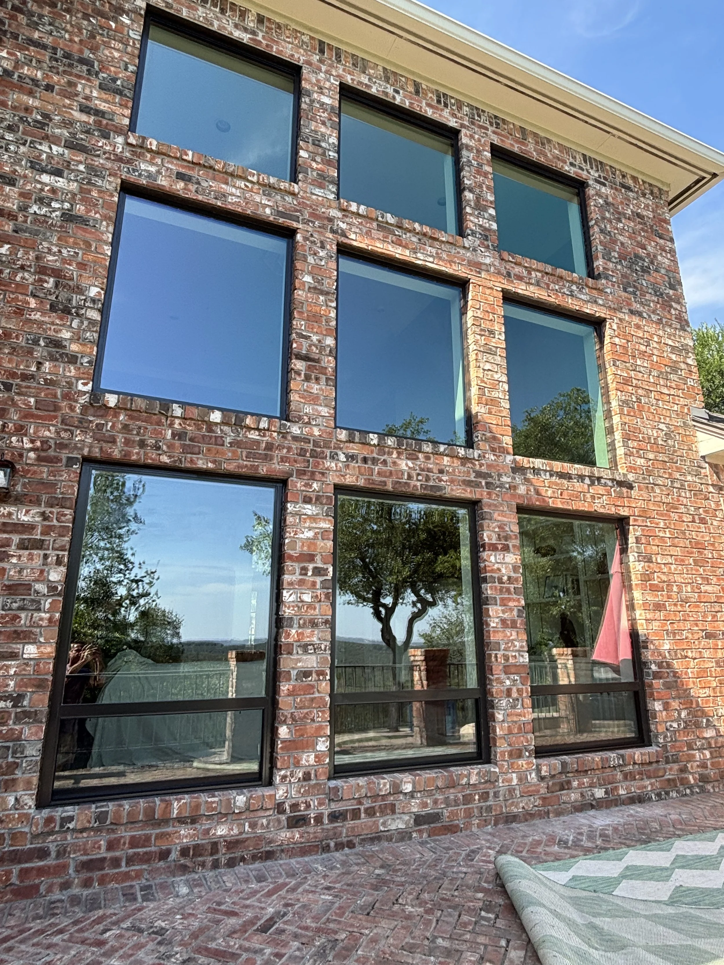 Exterior of a brick house with large, modern black-framed windows reflecting the sky, trees, and outdoor scenery, and a brick patio with a rolled-up green and white striped rug.