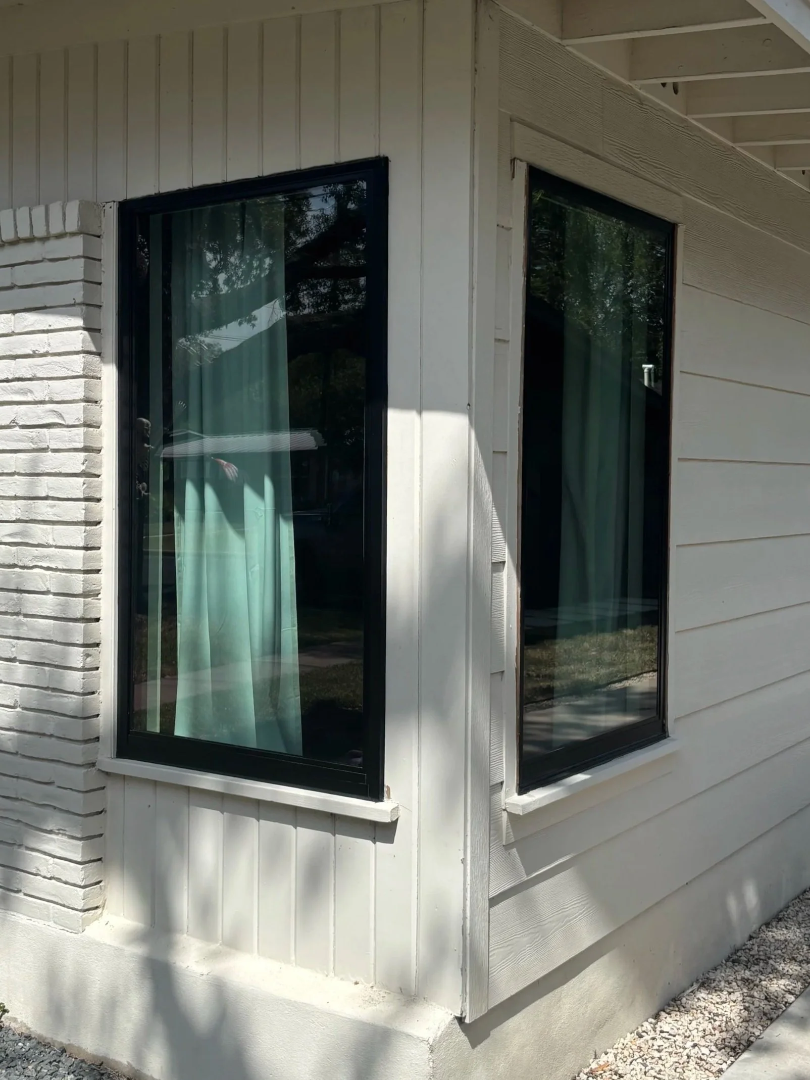 Corner of a white house with three windows featuring black frames and teal curtains, with reflections of trees and sky visible in the glass.