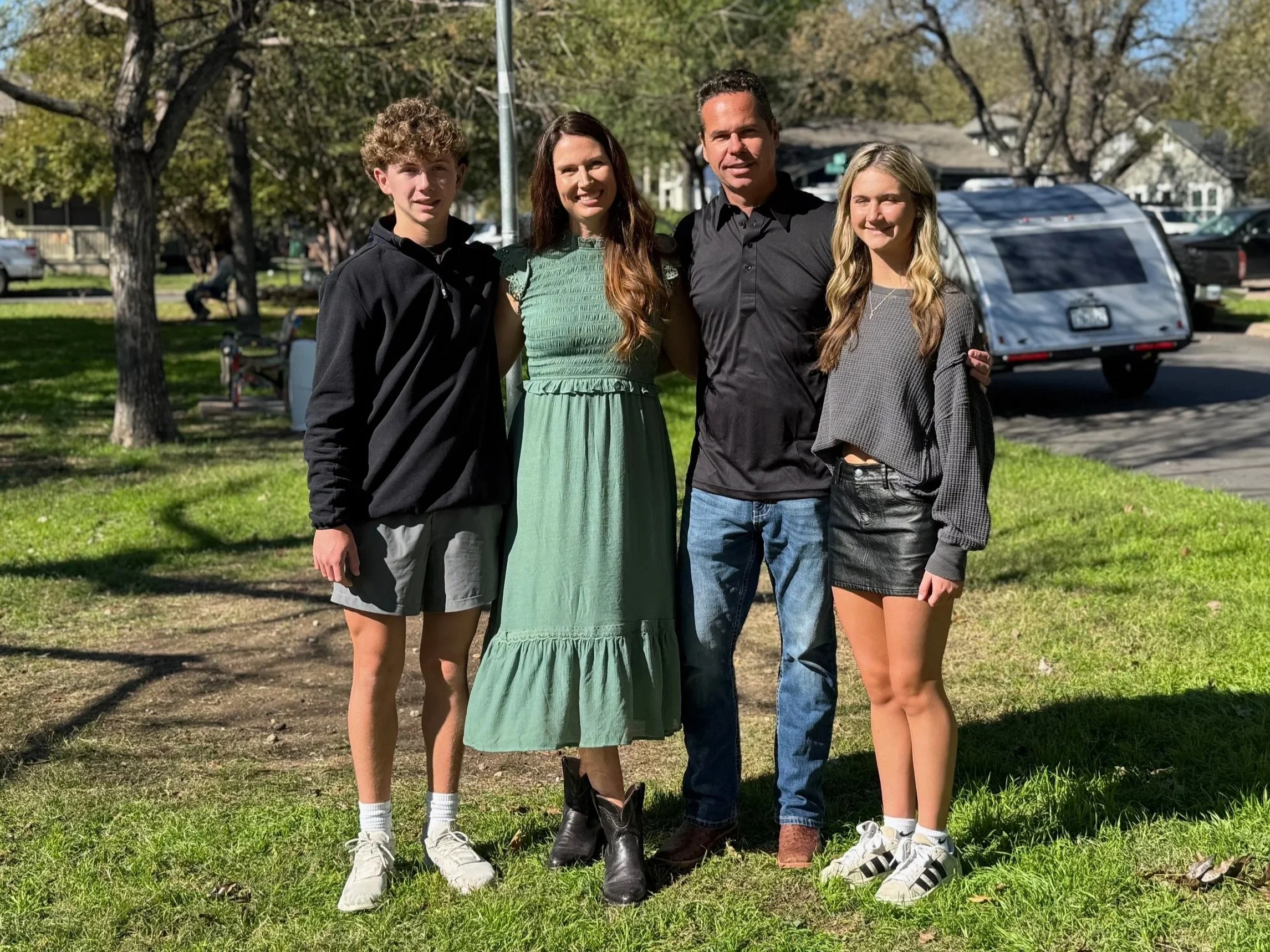 A family of four standing together outdoors on a sunny day, with trees, a small trailer, and parked cars in the background.