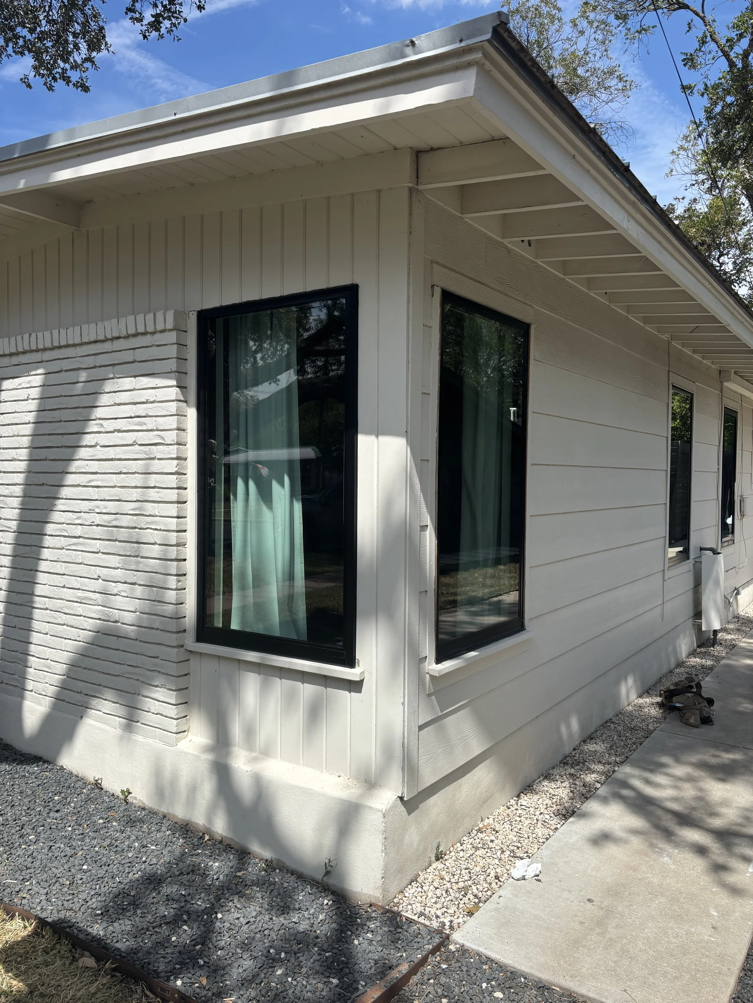 Side of a white house with large windows, exterior siding, and brickwork near a corner, with a sidewalk and gravel at the base.