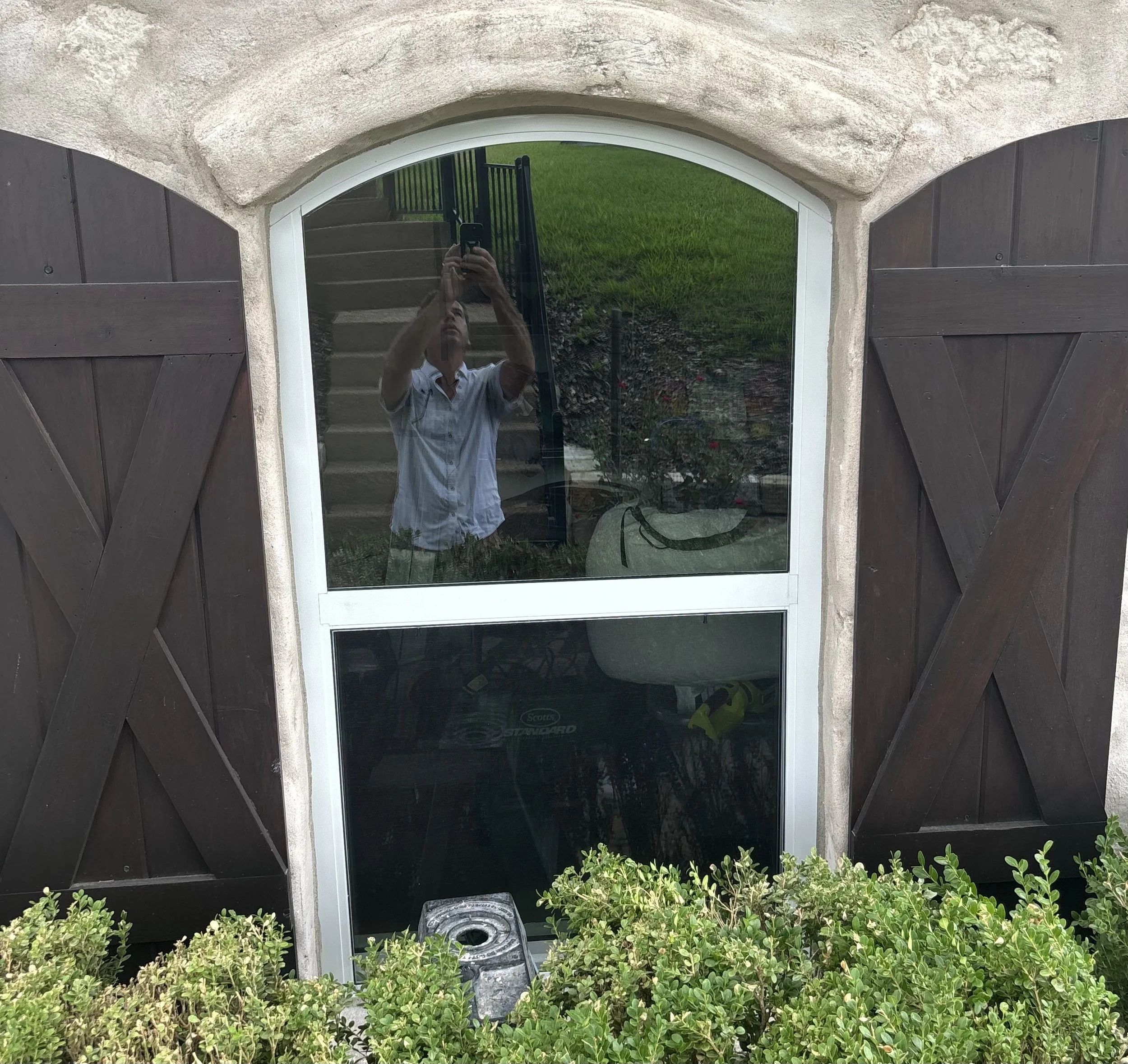 Reflection of a man taking a photo of a window with wooden shutters, with outdoor grass, stairs, and bushes visible.