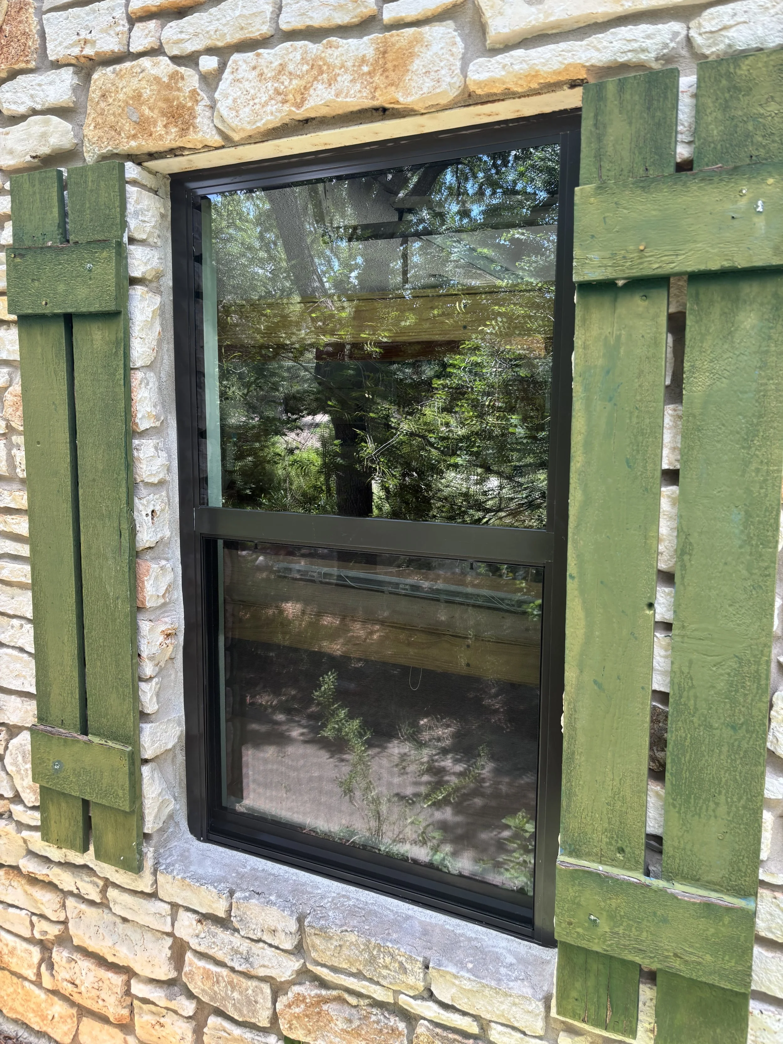 A window with black metal frame, reflecting trees and sky outside, set in a stone wall with green wooden shutters.