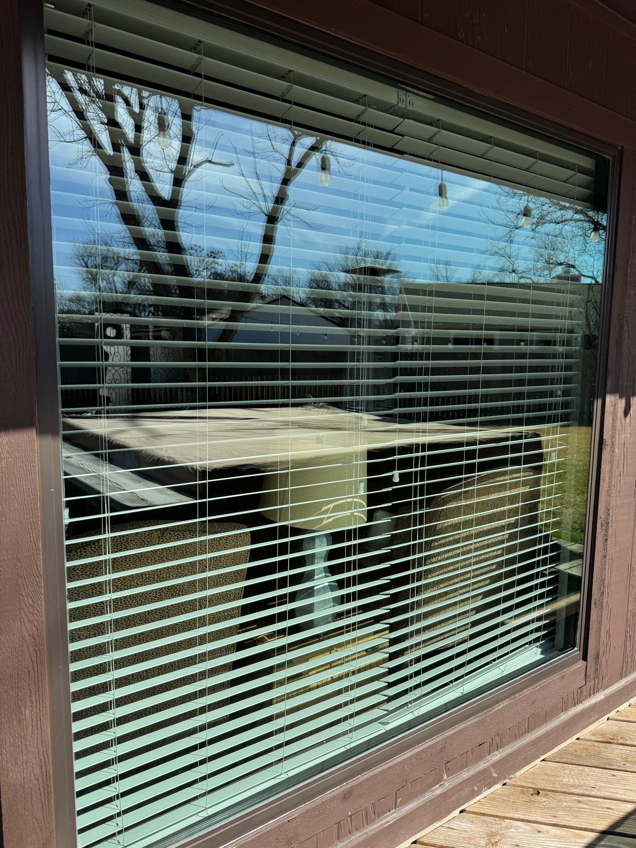 View through window with blinds showing outdoor furniture and a view of trees and houses in the background.