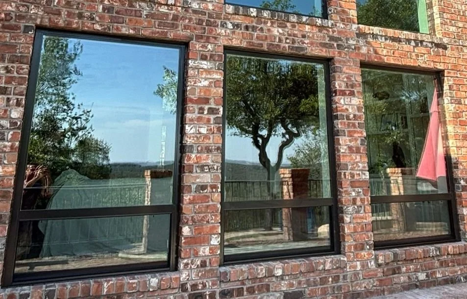 Close-up of three large windows in a brick building, reflecting trees and a clear sky outside.
